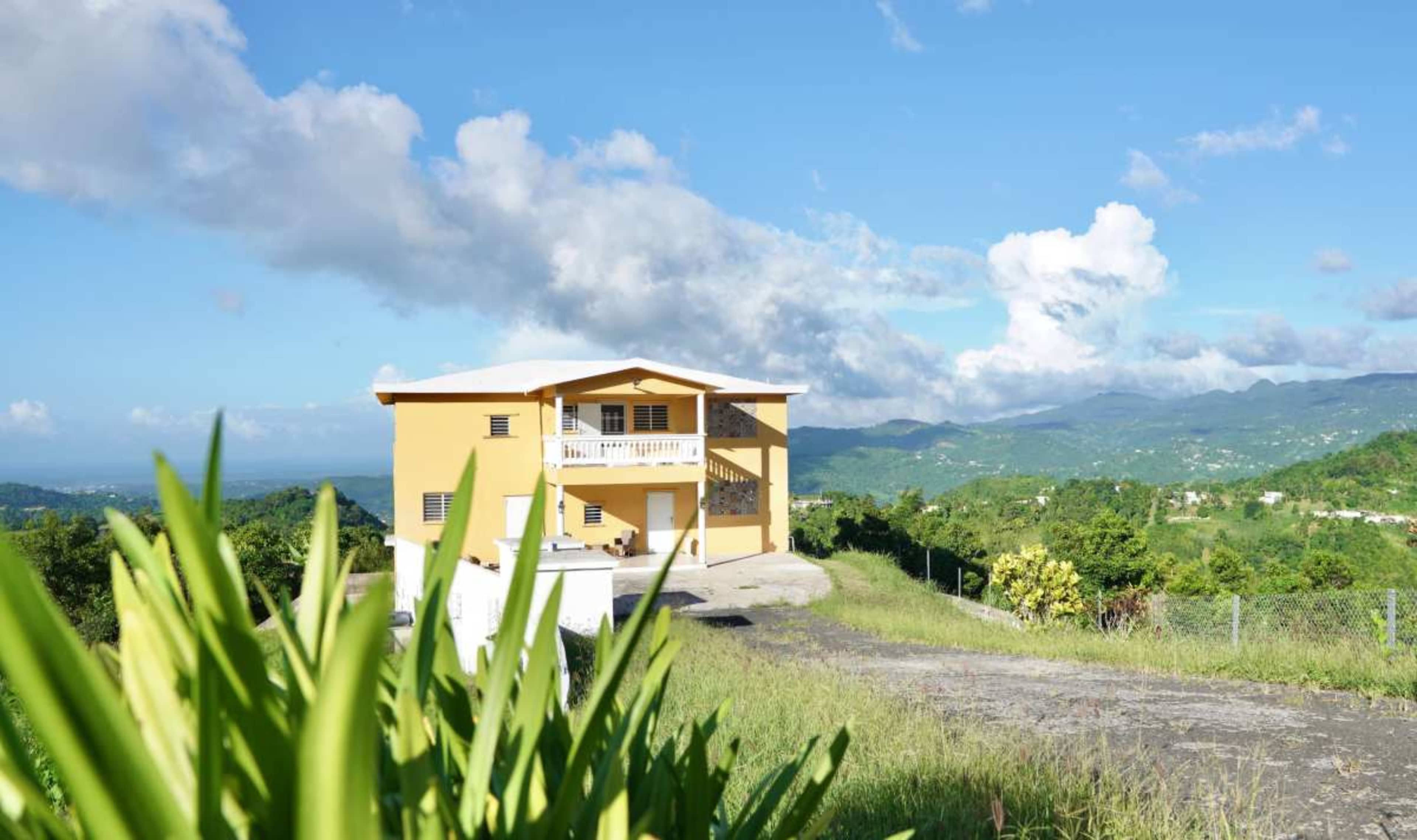 A yellow house with a balcony overlooks a green landscape and distant mountains under a partly cloudy sky.