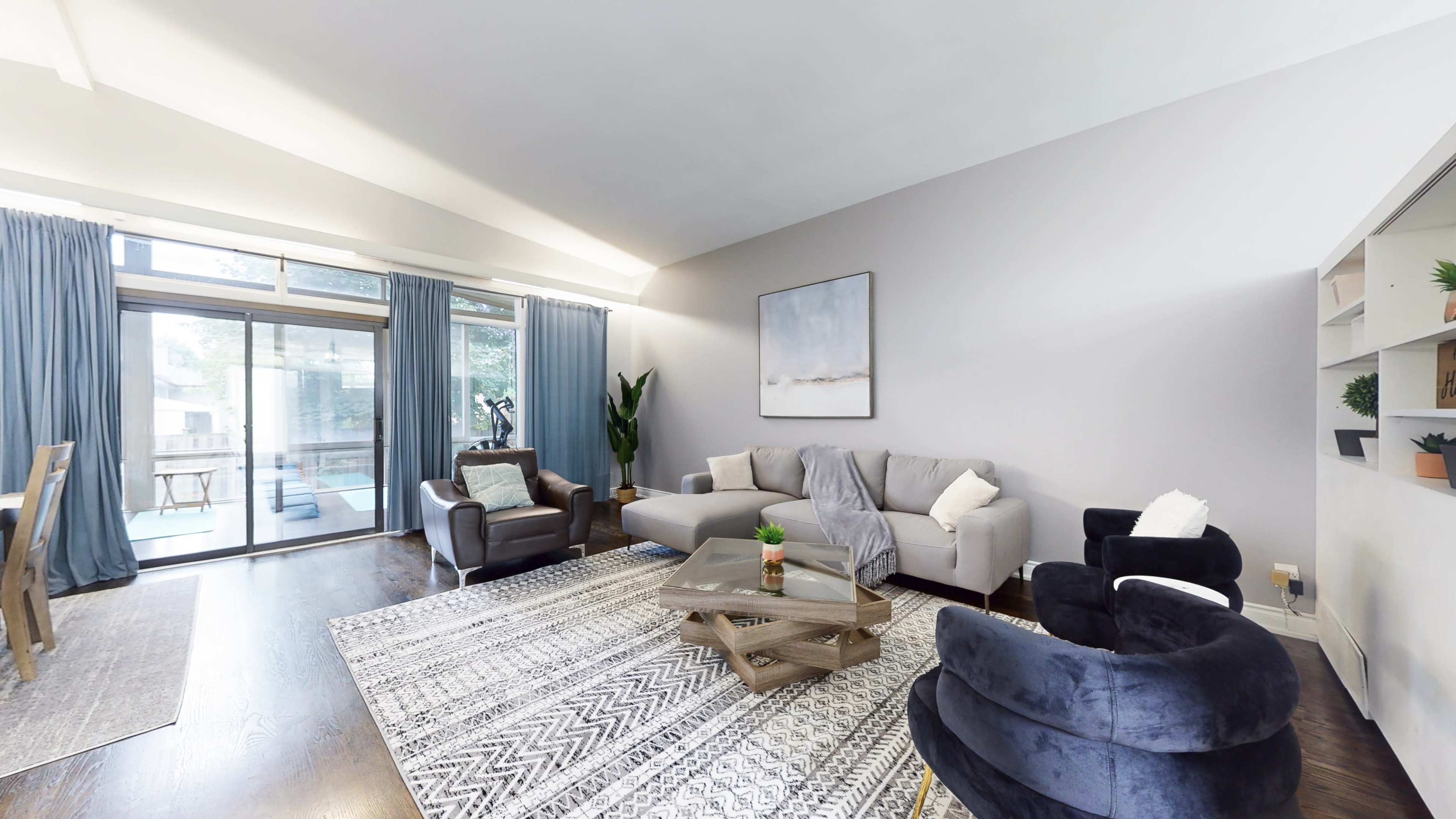 The living room features a light gray sofa, two dark chairs, a coffee table, and large windows with sheer curtains letting in natural light.