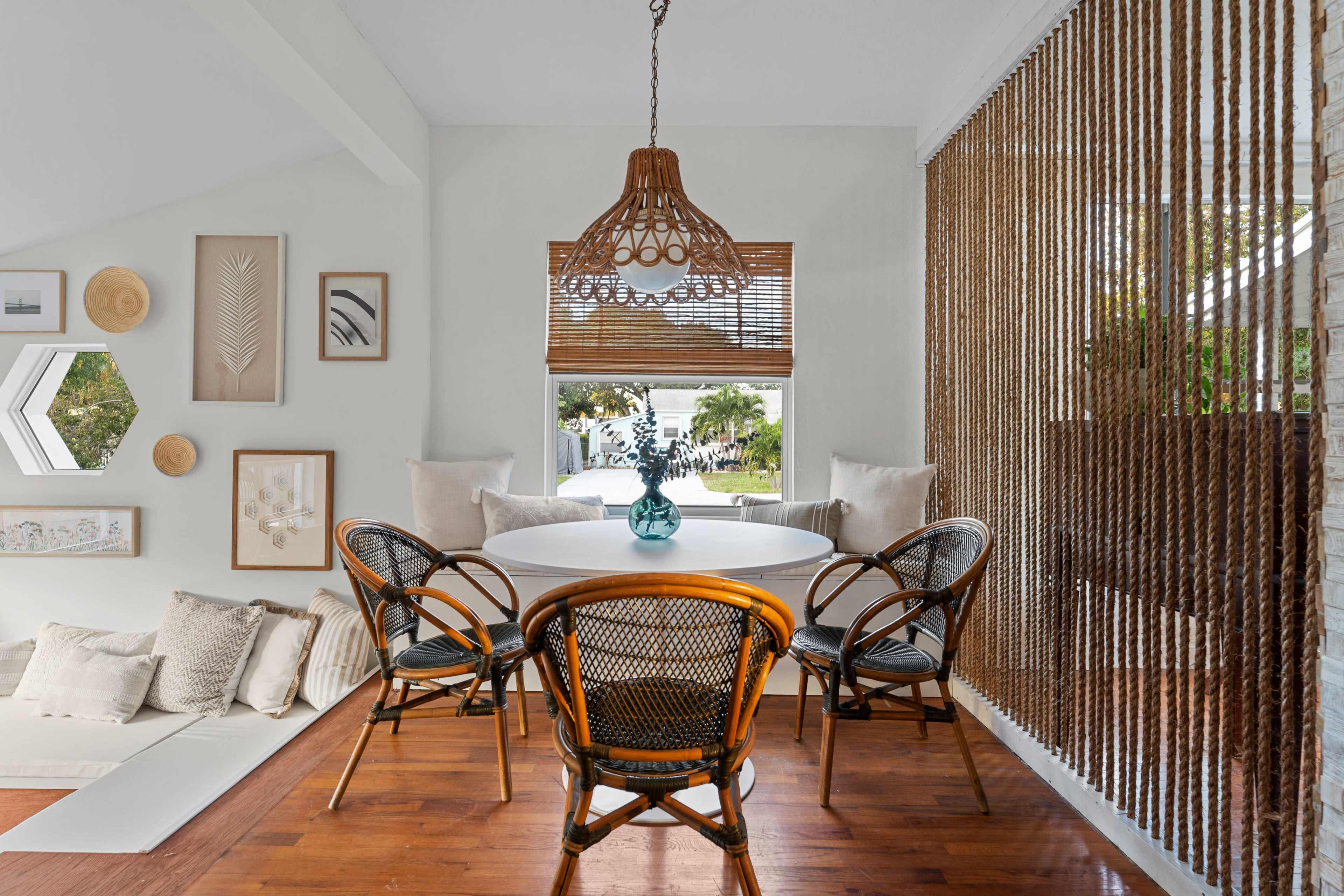 A dining area features a round table surrounded by four black wicker chairs, with a large window offering a view of the outdoors and light-colored walls adorned with framed artwork.