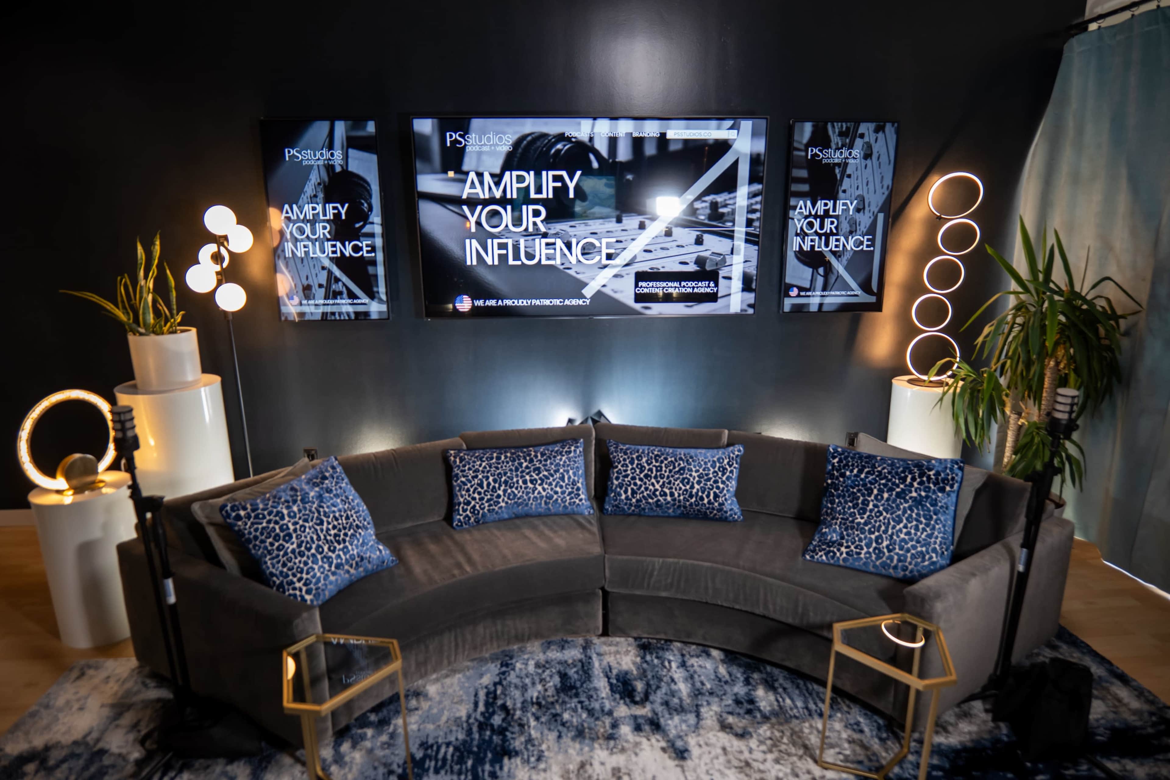 The image shows a modern lounge area featuring a curved gray sofa with decorative cushions, alongside three large screens displaying the text "AMPLIFY YOUR INFLUENCE," and plants in white pots.