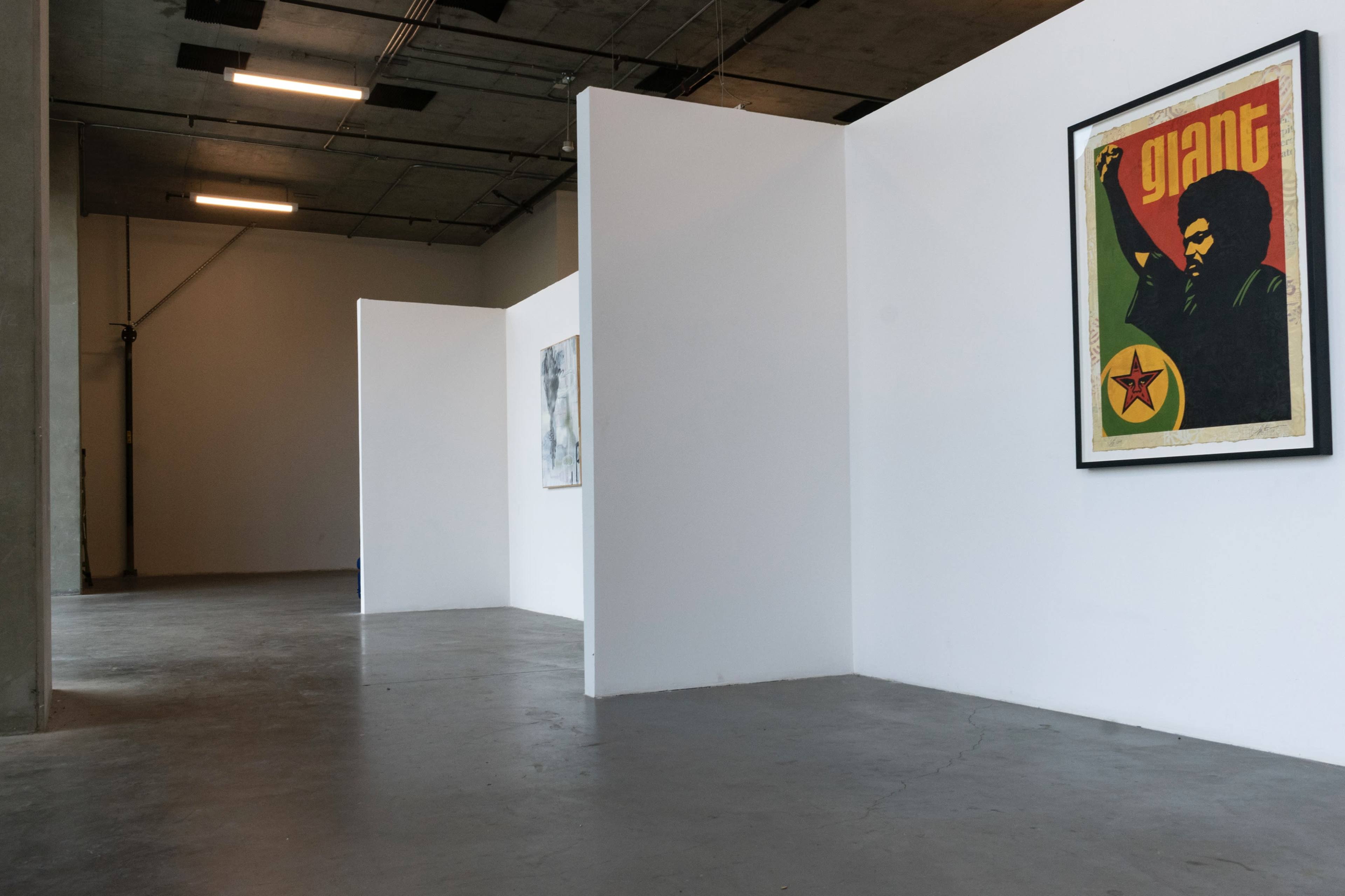 A spacious gallery with white walls and a single framed poster on the right side featuring a figure raising a fist, while additional artworks are partially visible behind the walls.