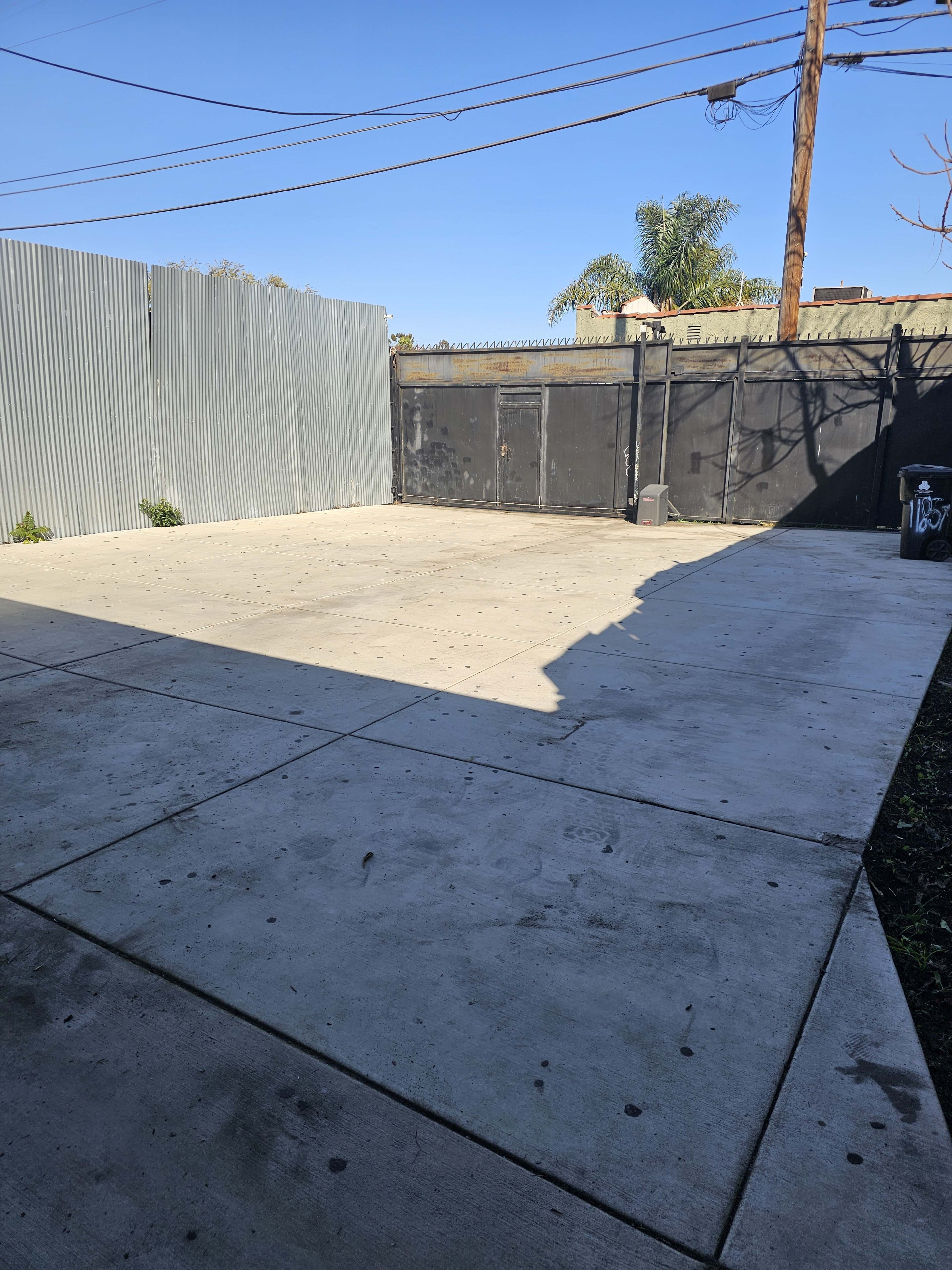 The image shows an empty, concrete enclosure with a corrugated metal fence and a black gate, under a clear blue sky.