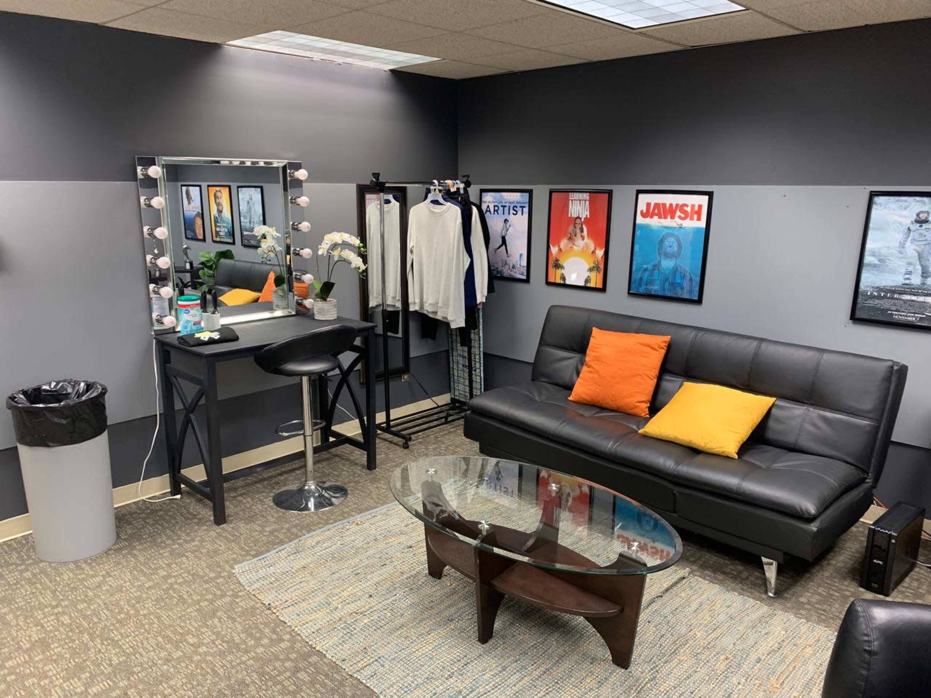 A small lounge area featuring a black sofa with orange pillows, a glass coffee table, a dressing table with lights, and framed movie posters on the walls.