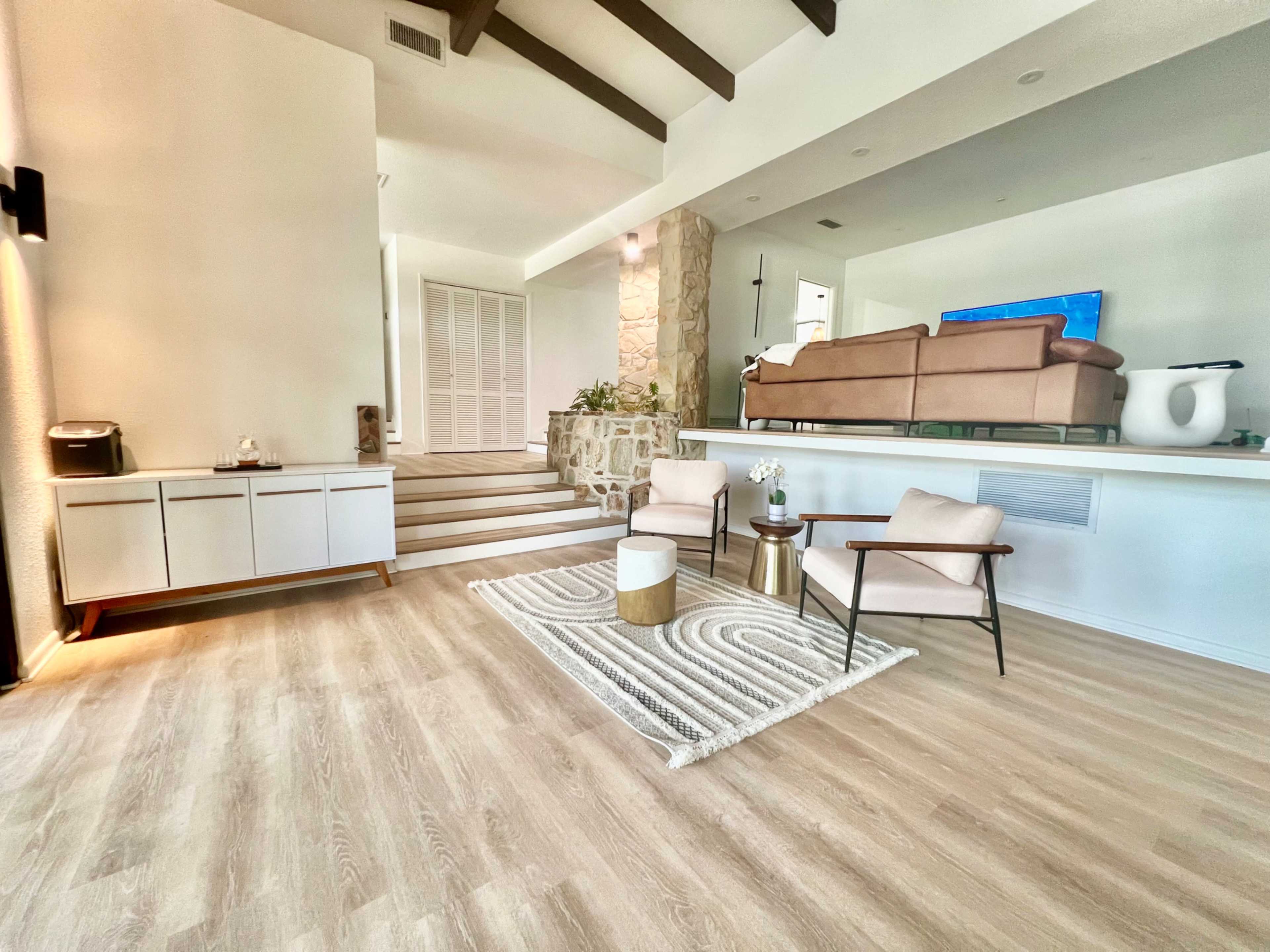 The interior space features a light wood floor with a seating area comprising two chairs and a decorative rug, alongside a stone wall and steps leading to a separate section with a large sofa and television.