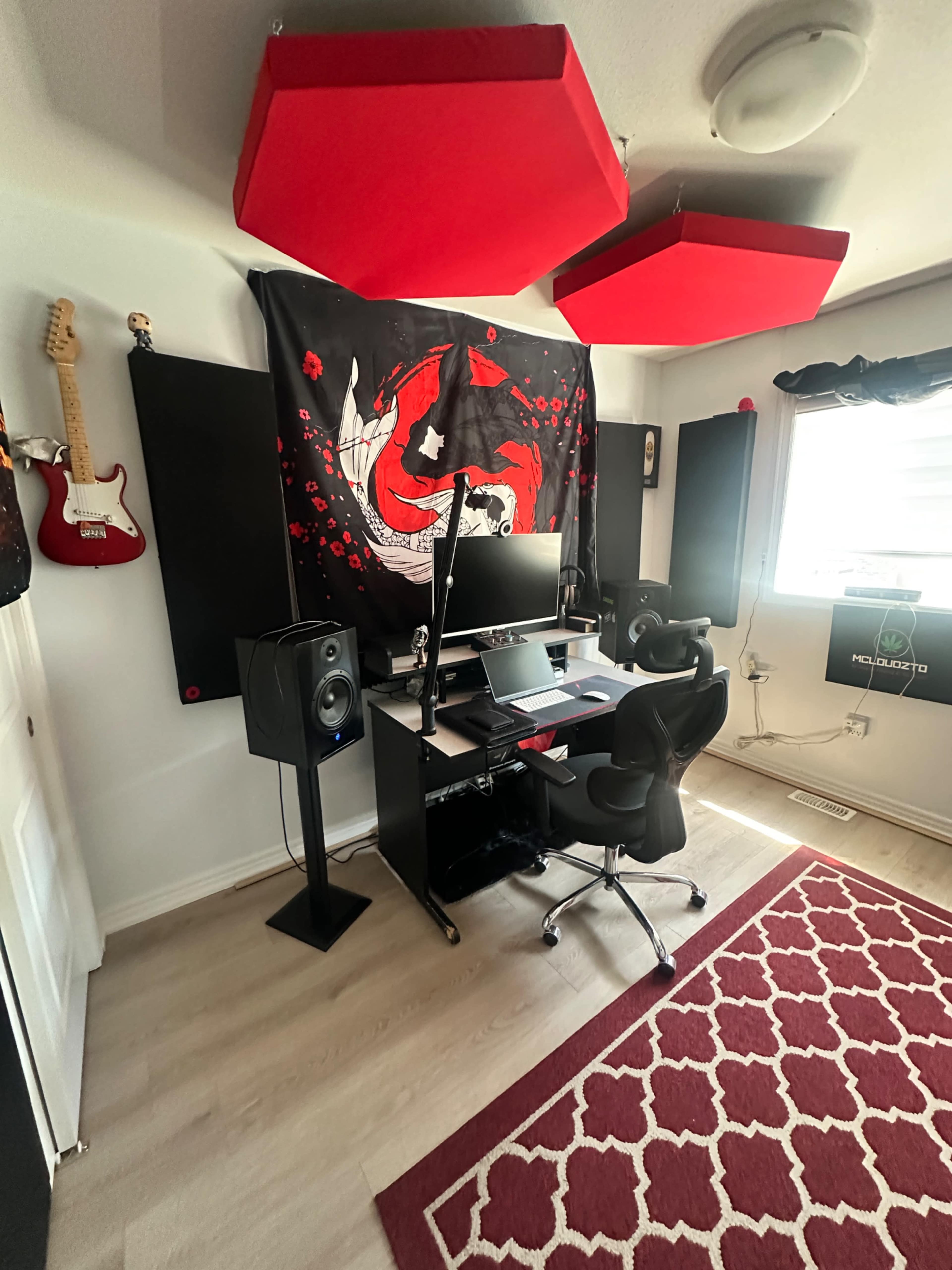 The image shows a home studio setup featuring a black desk with a computer, two speaker monitors, and acoustic panels on the walls, accented by red and black decor.