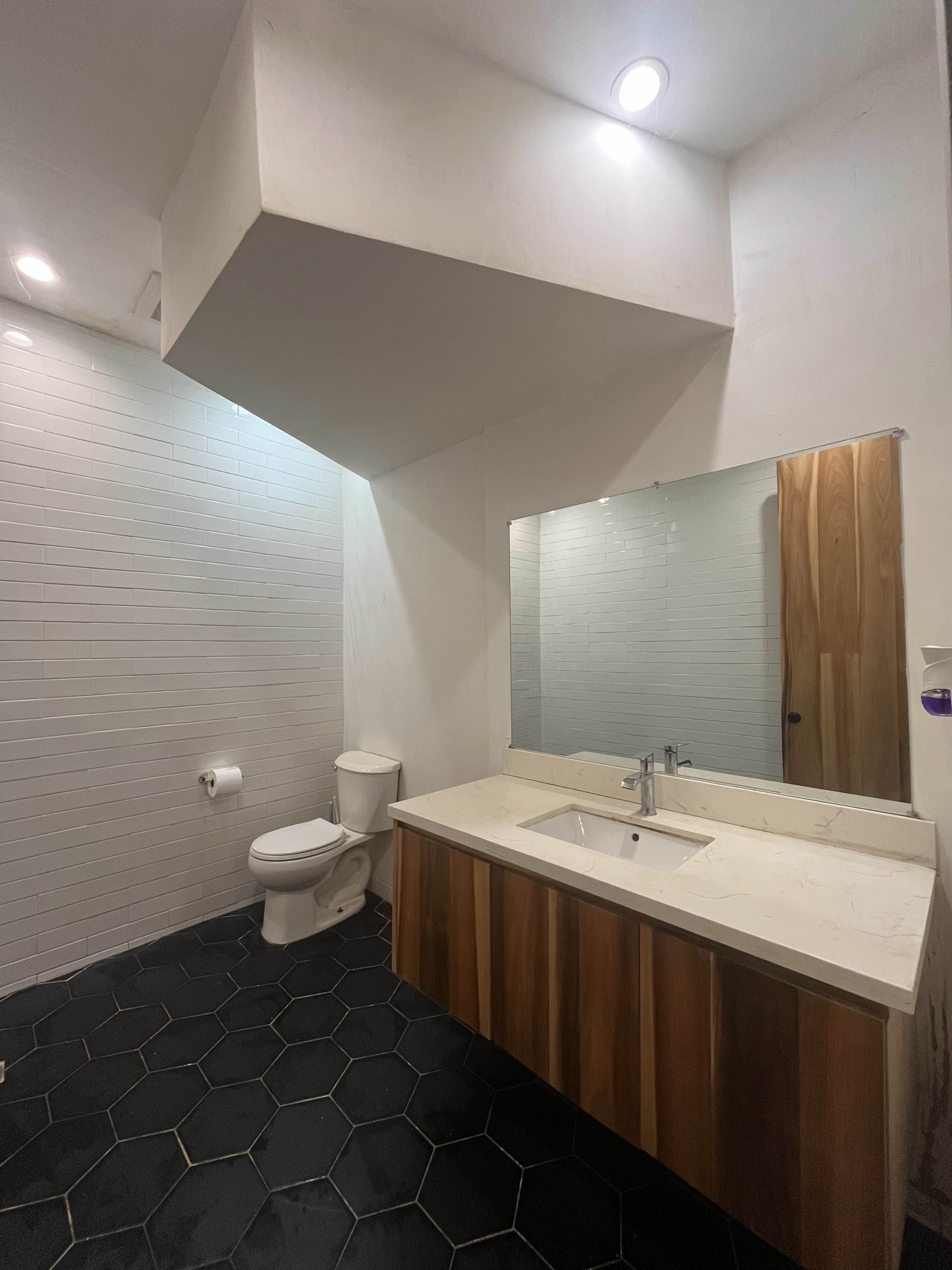 The image shows a modern bathroom featuring a wall-mounted sink with a wooden cabinet below, a large mirror, a toilet, and hexagonal floor tiles.