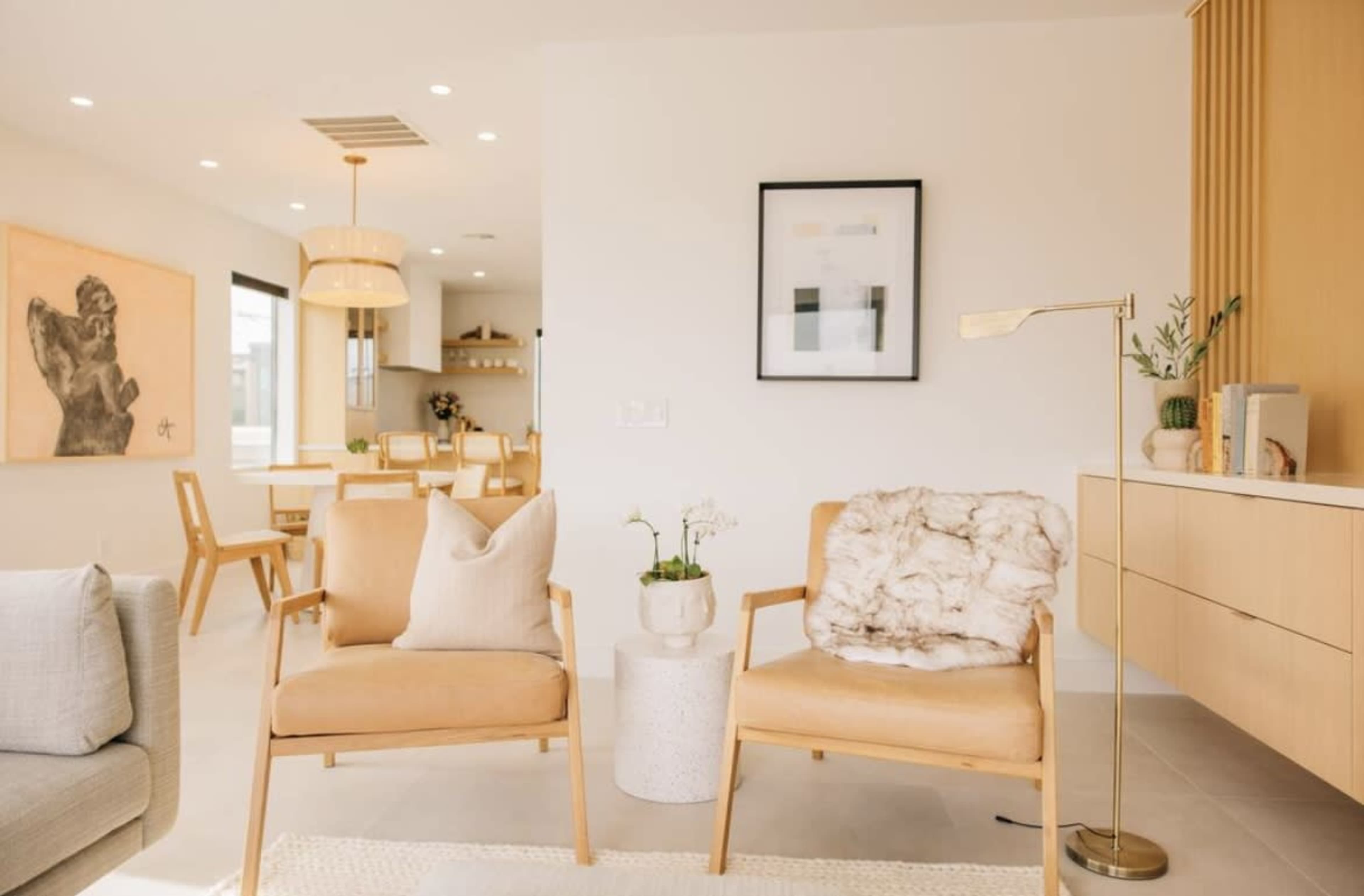 A modern living room with two light brown armchairs, a small side table, and a minimalist decor featuring a wall art piece and a dining area in the background.