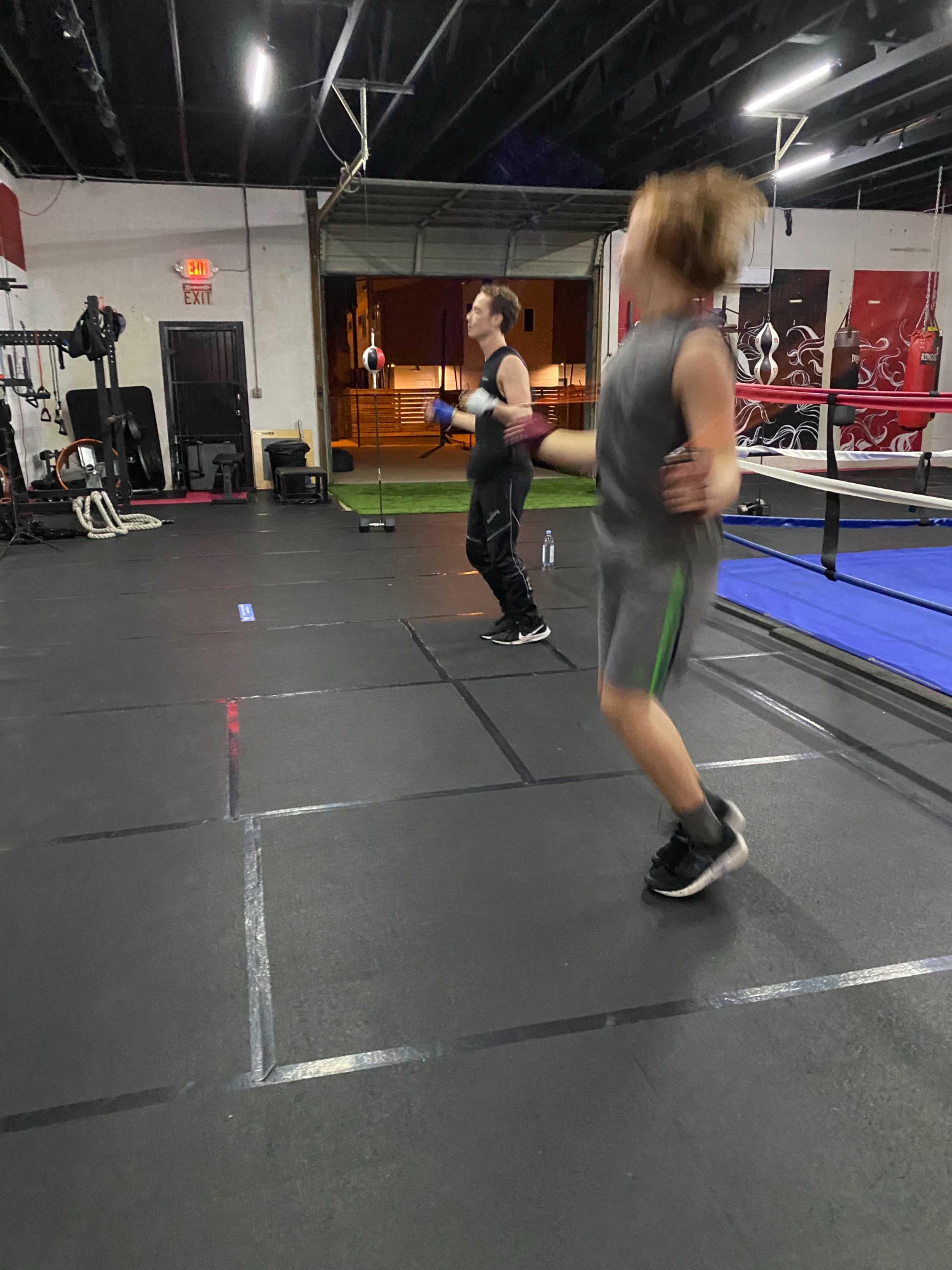 Two individuals are jumping rope in a gym with workout equipment and a boxing ring in the background.