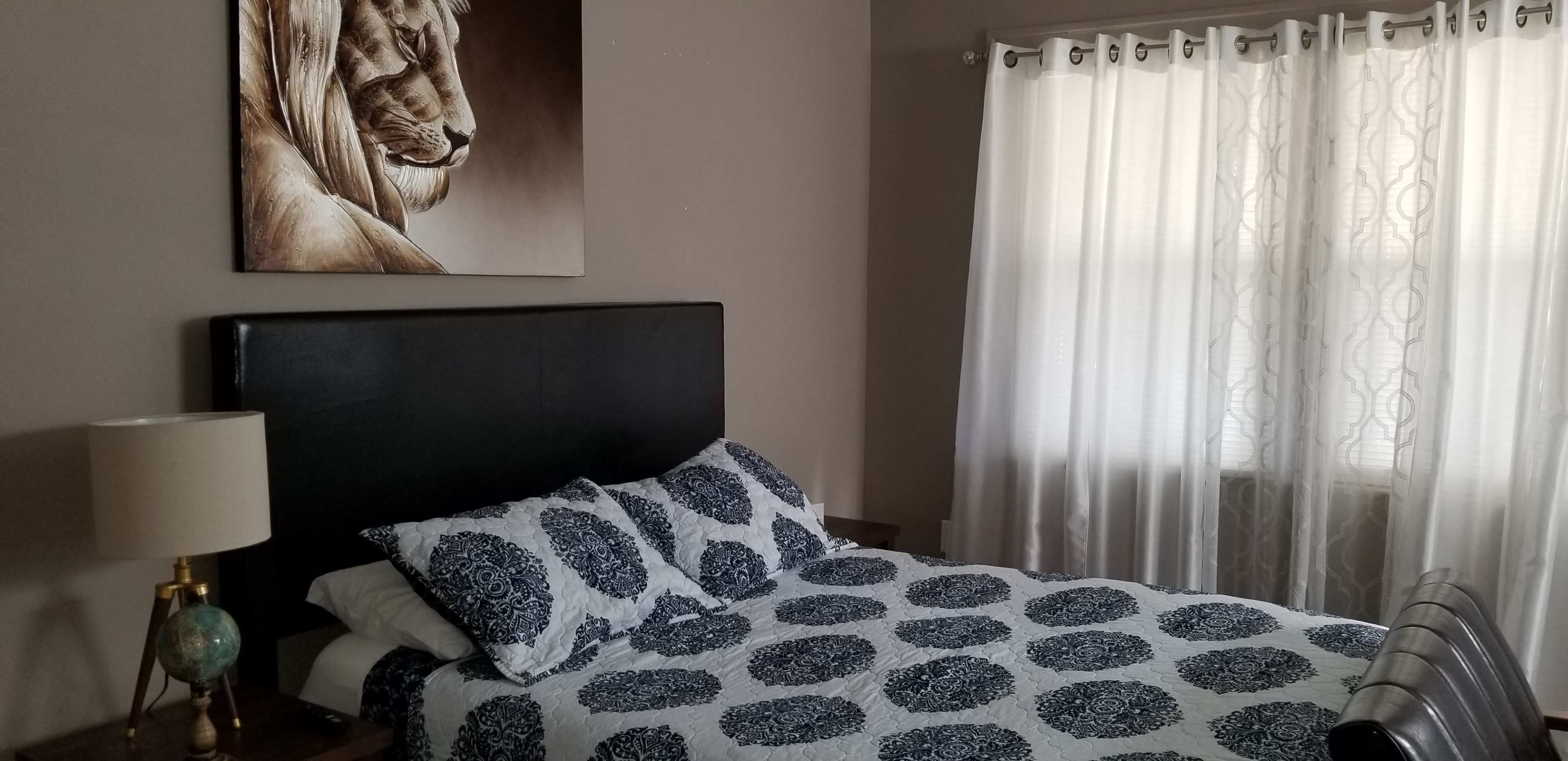 A bedroom features a bed with a patterned blue and white bedspread, a lion portrait on the wall, and sheer white curtains allowing light to enter.