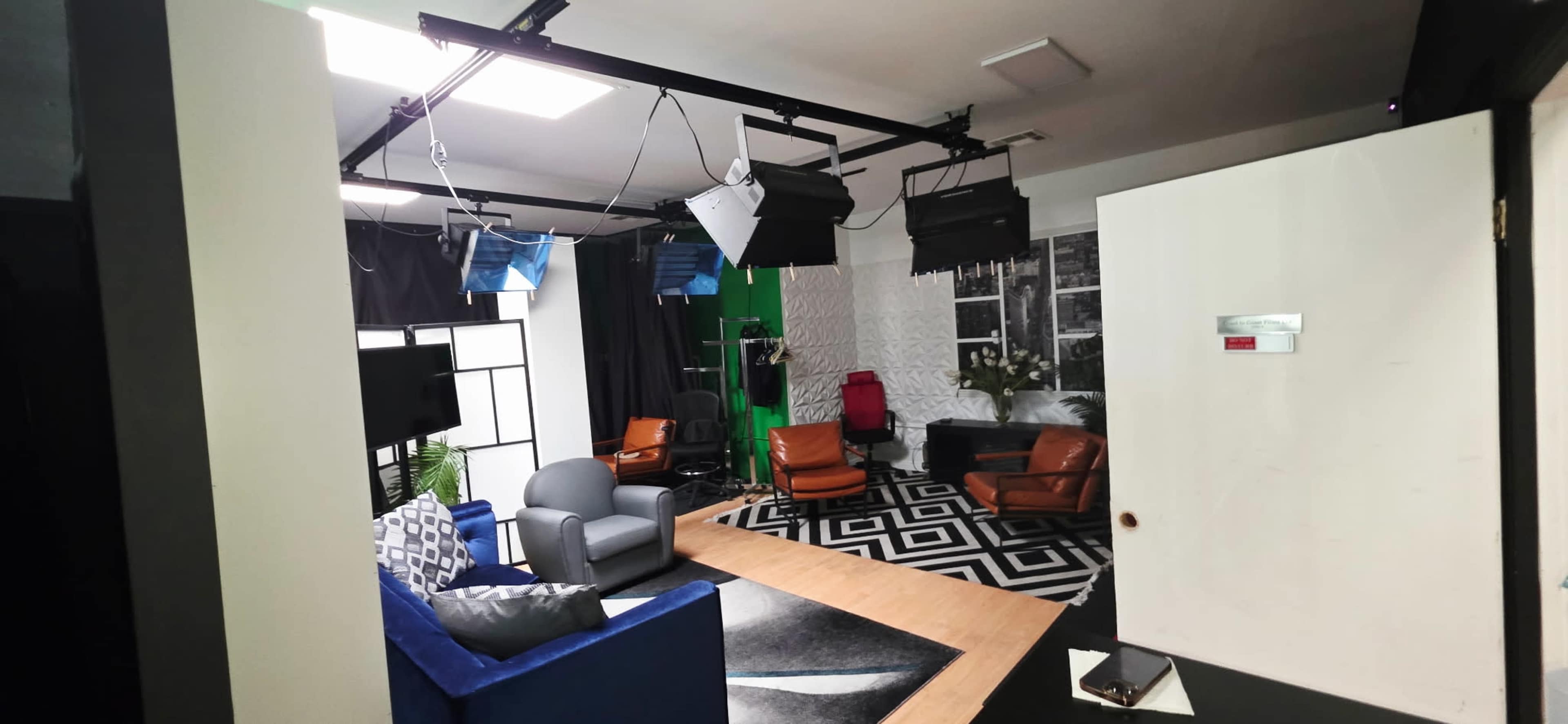 The image shows a well-lit studio setup with various seating areas, including a blue couch and brown chairs, alongside lighting equipment and a green backdrop.