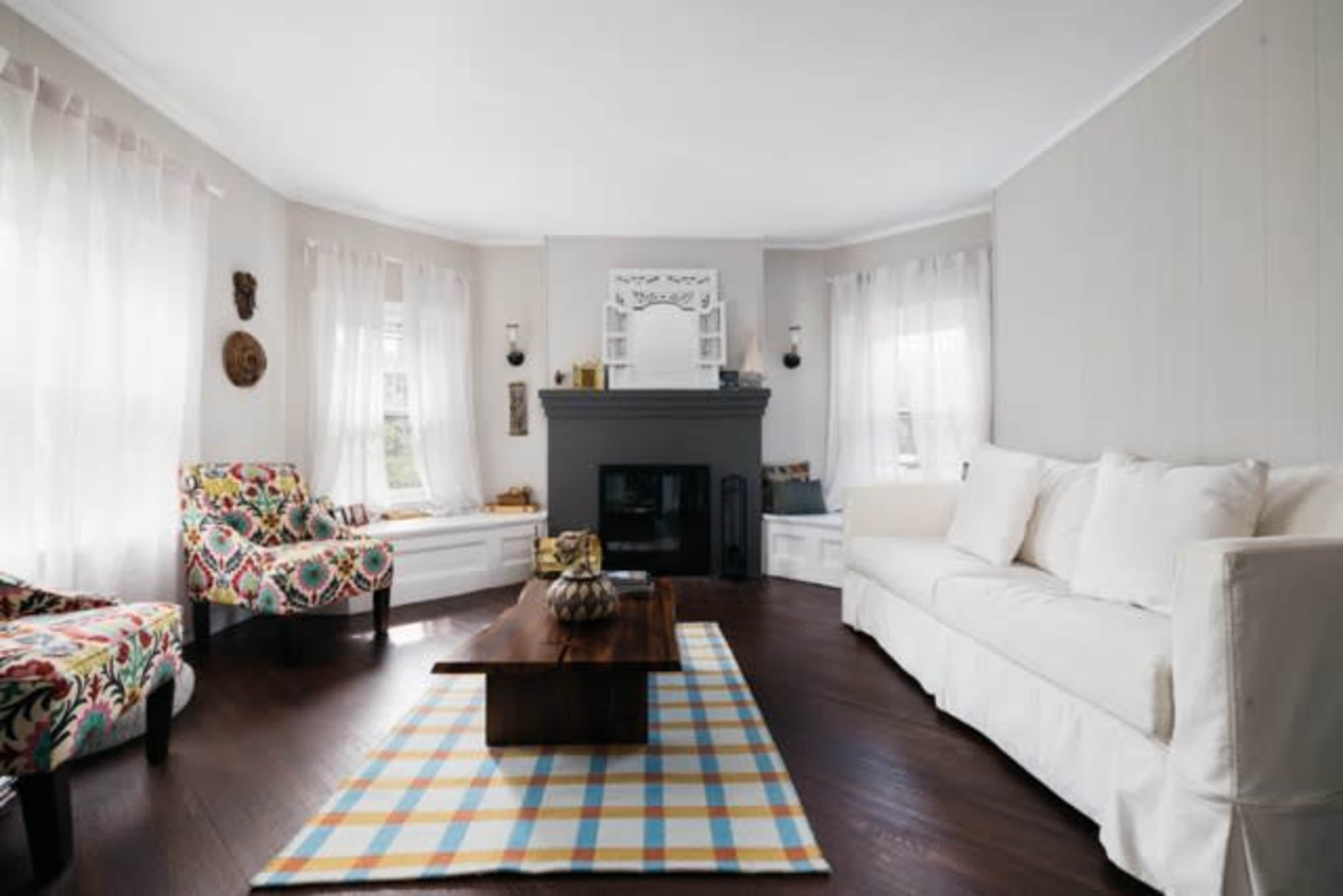 A cozy living room features a white sofa, colorful patterned chairs, a dark wood coffee table, and large windows with sheer curtains.