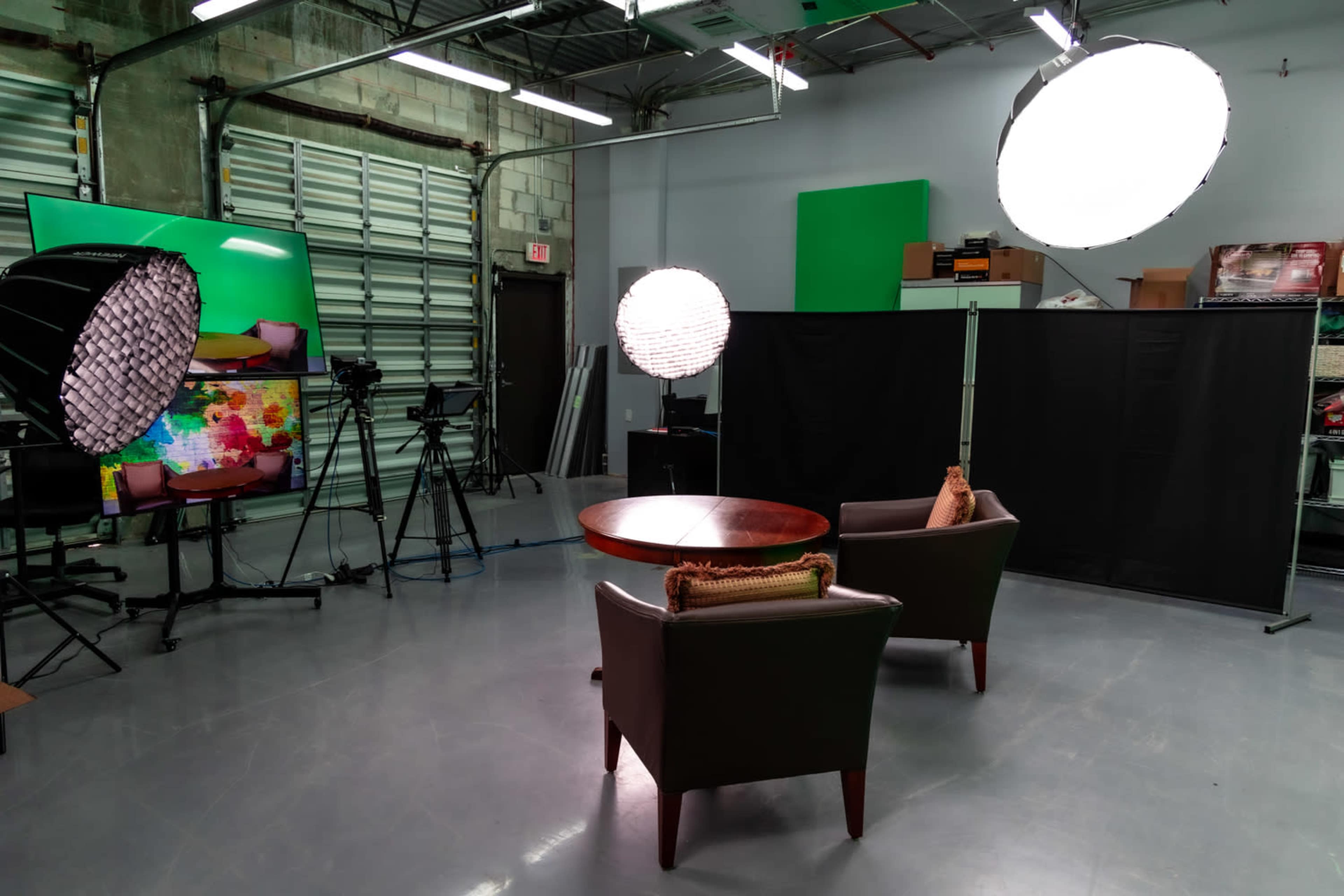 The image shows a video production studio with two chairs positioned near a round table, surrounded by lighting equipment and a green backdrop.