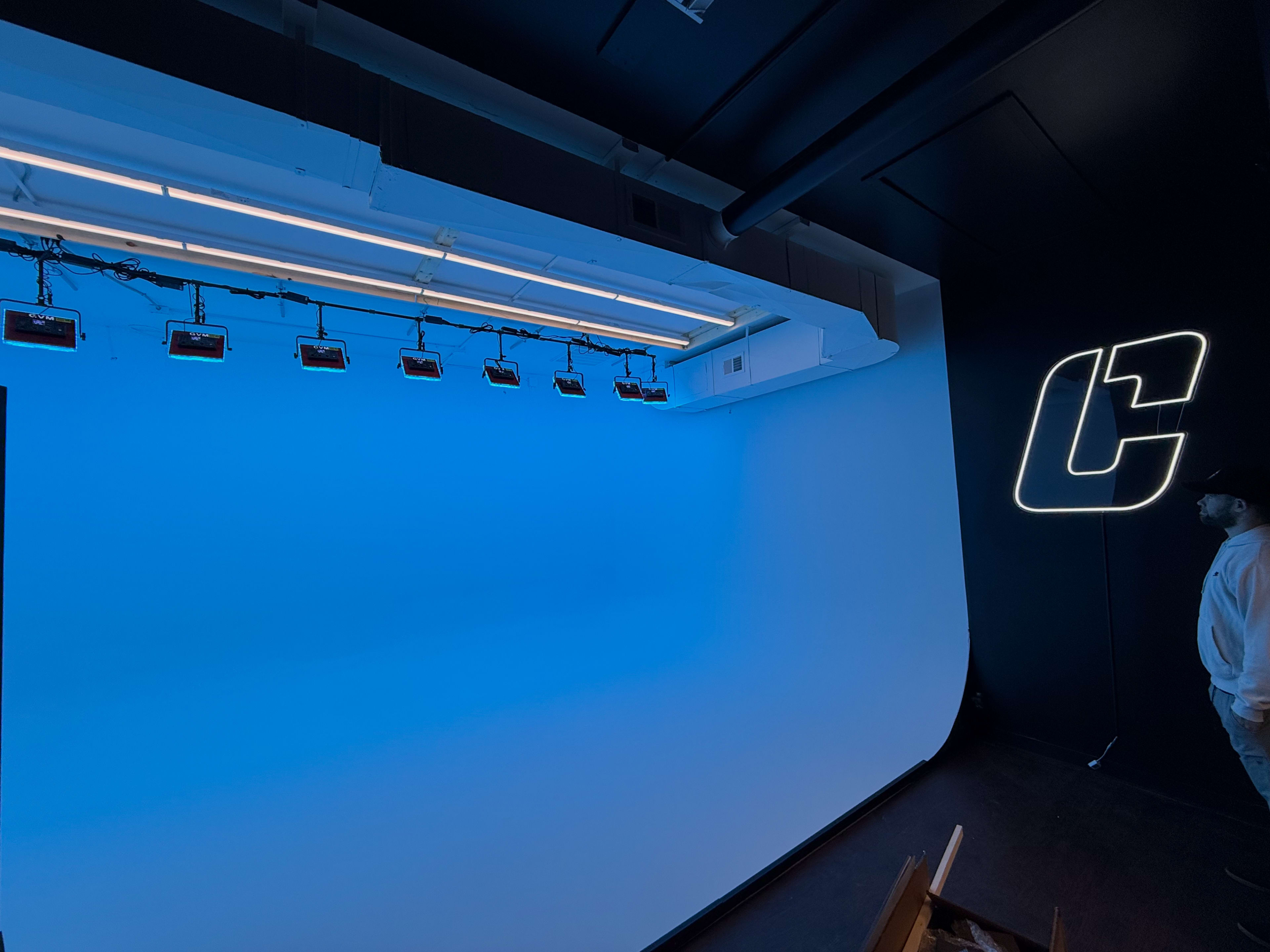 The image shows a studio space with a blue backdrop, lighting fixtures mounted on the ceiling, and a neon sign on the wall.