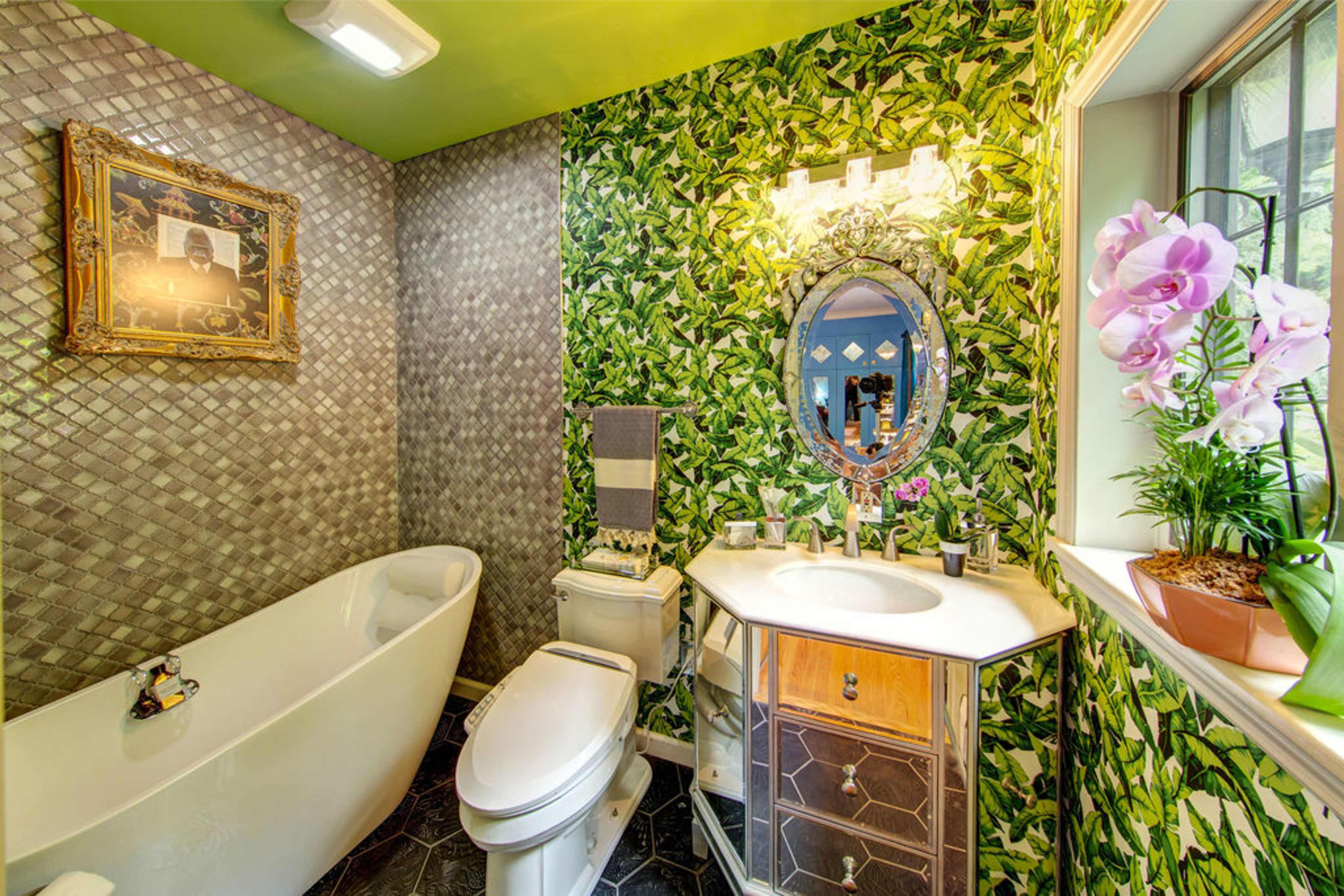 The image shows a small bathroom featuring a freestanding bathtub, a modern vanity with a mirror, and leafy wallpaper accented by a decorative light fixture.