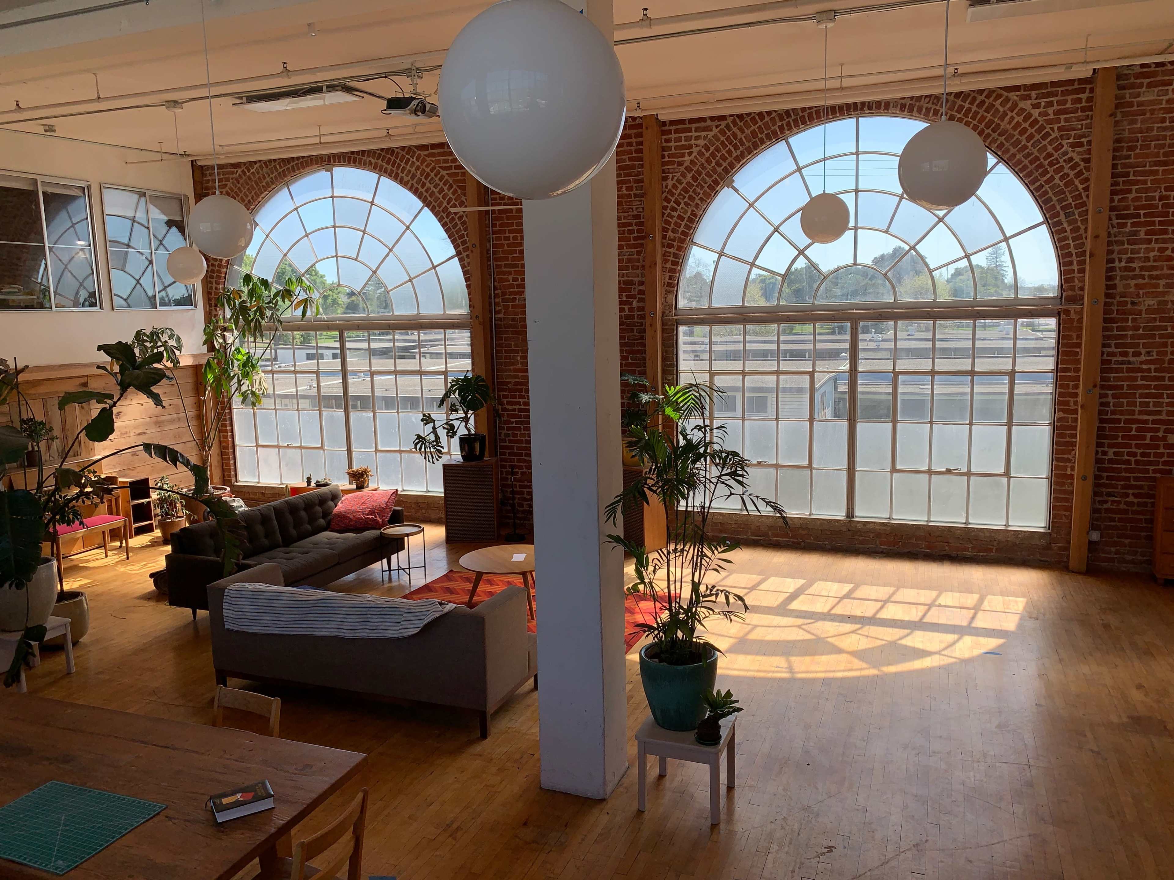 The image shows a spacious loft with large arched windows, brick walls, and a seating area featuring a couch, plants, and a coffee table.