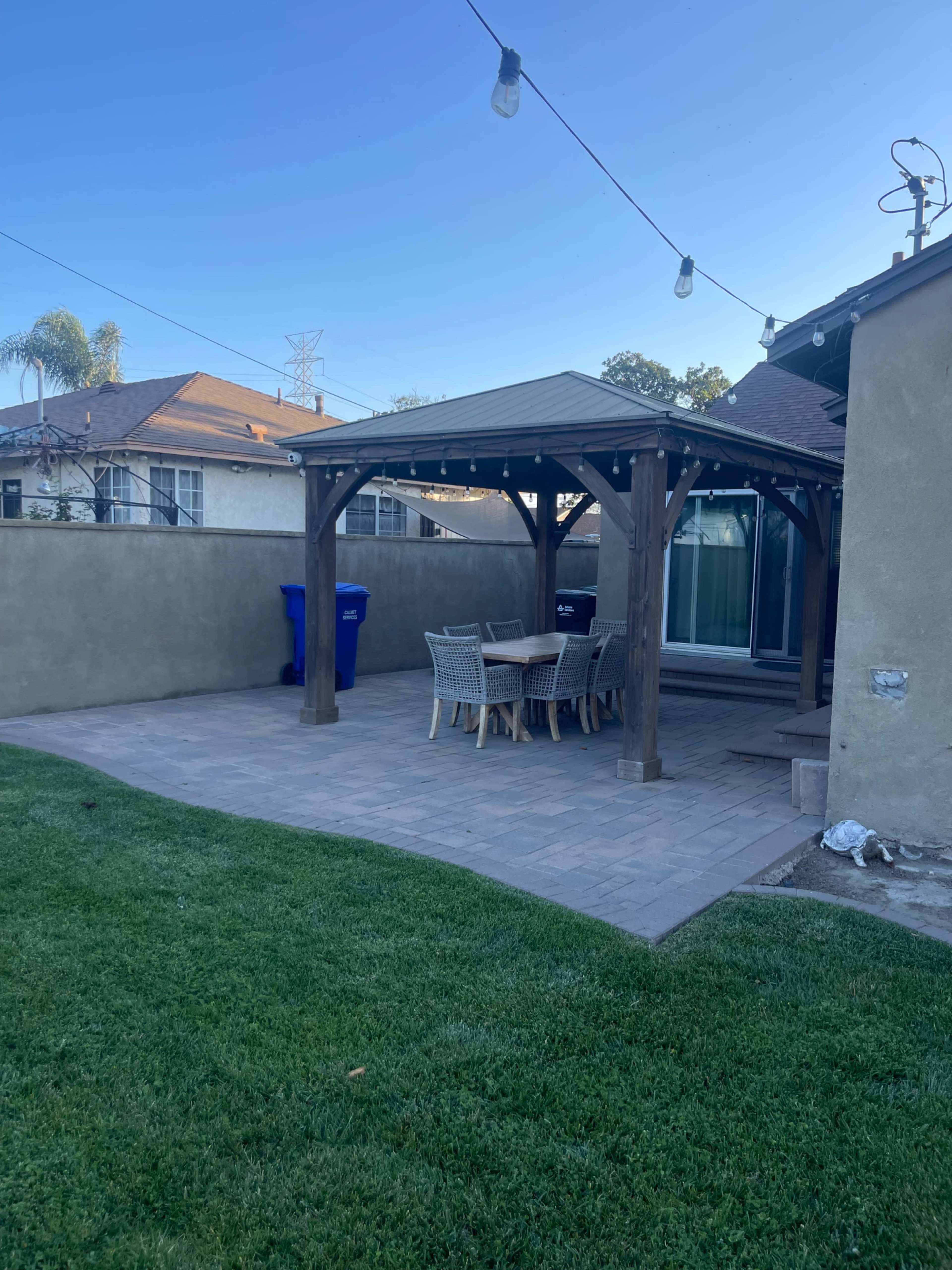 The image shows a backyard patio area with a shaded wooden gazebo, a table surrounded by chairs, and green grass.