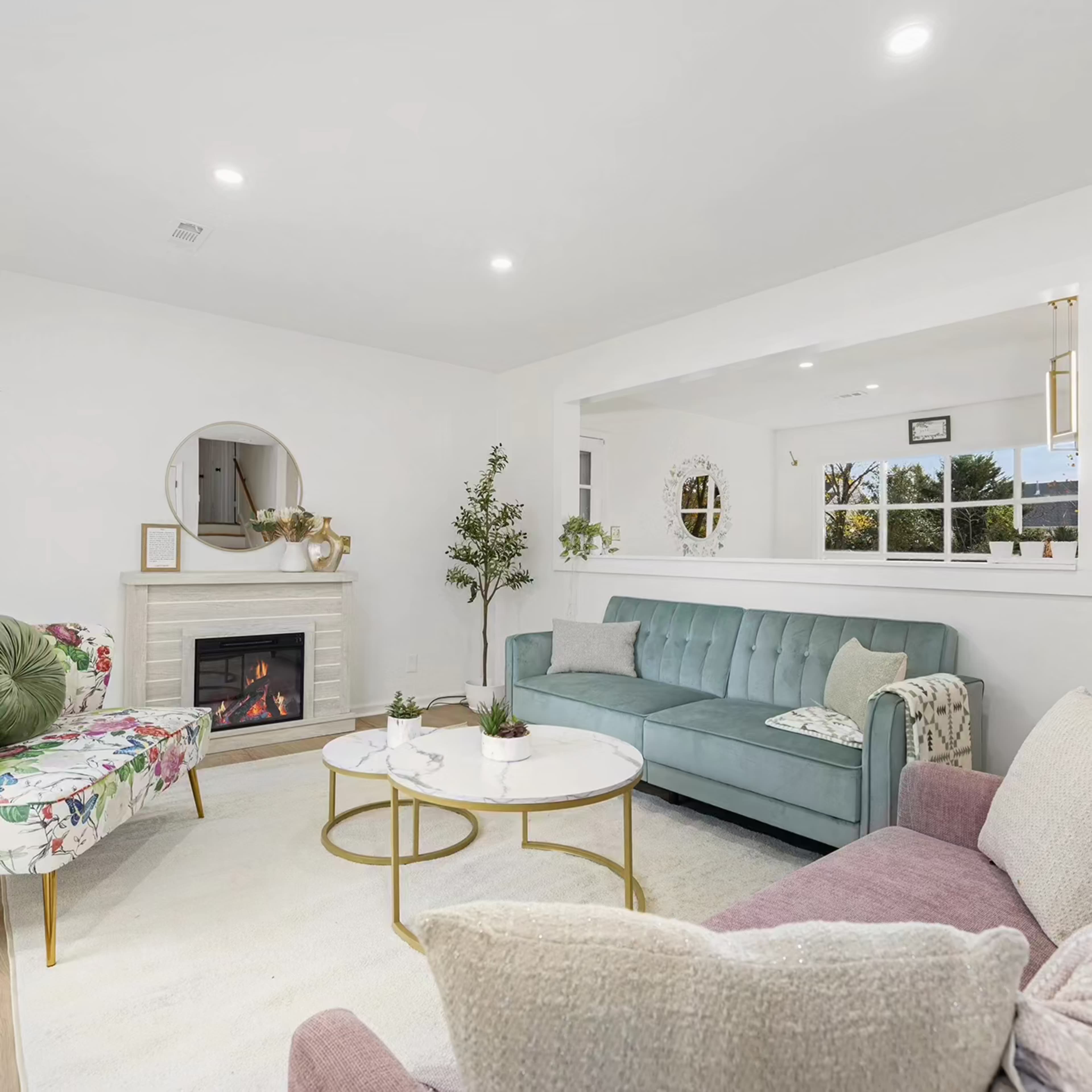 A bright living room featuring a teal sofa, a pink armchair, a floral accent chair, a white rug, and a fireplace with a round mirror above it.
