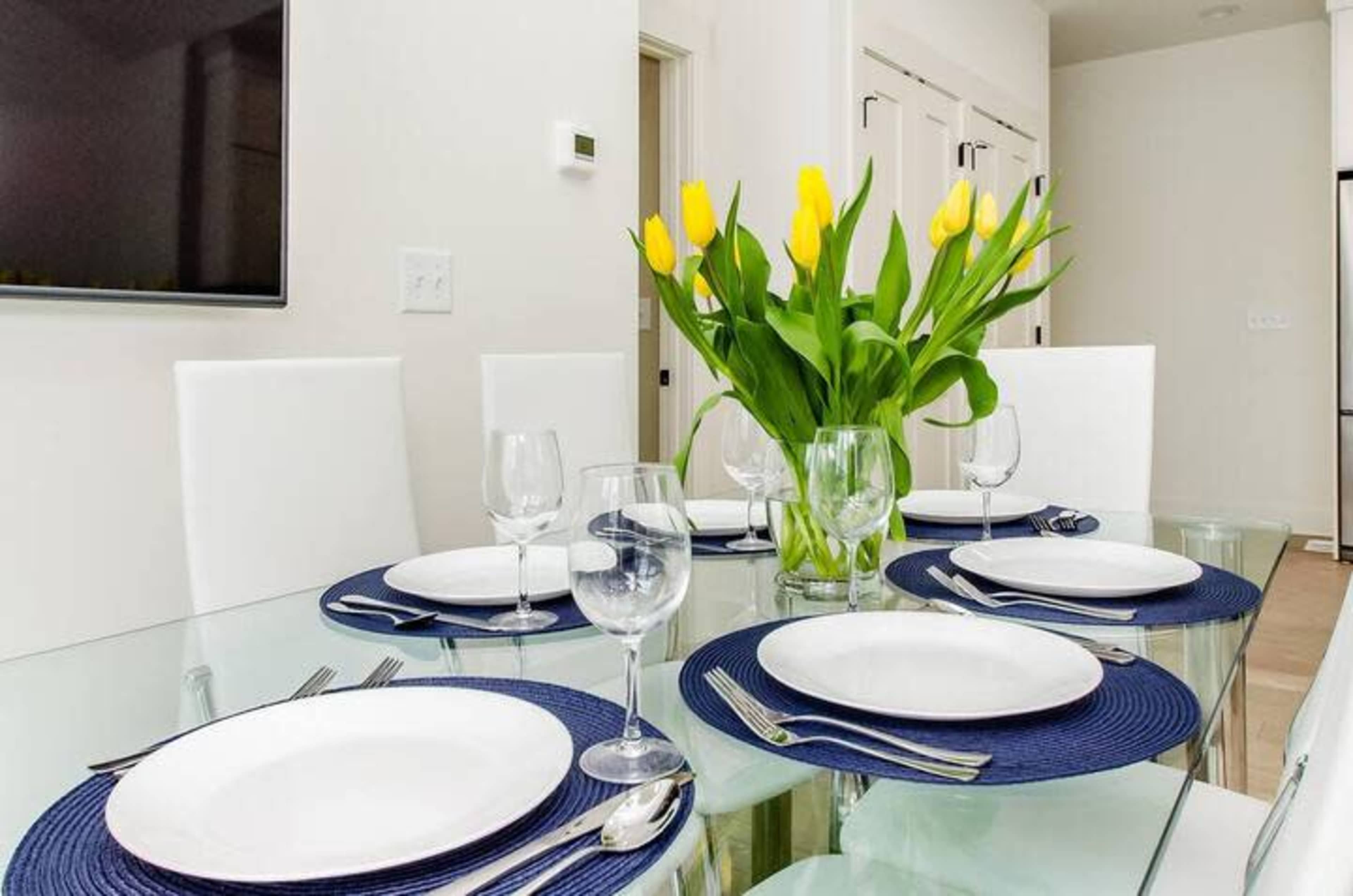 A glass dining table is set with white plates and cutlery, surrounded by white chairs, and features a vase of yellow tulips at the center.
