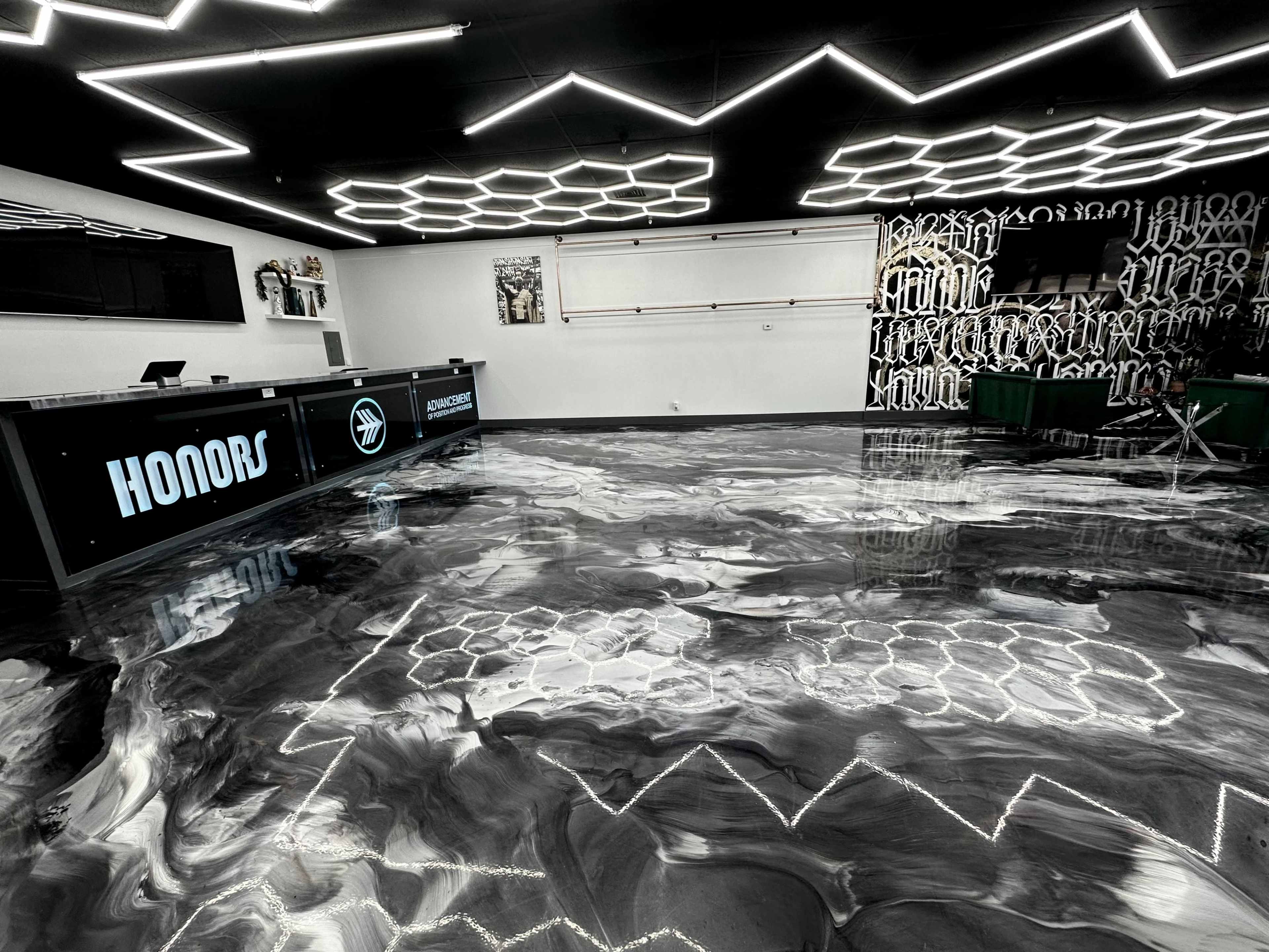 A modern interior space with a polished black and white floor, illuminated geometric ceiling lights, and a counter labeled "HONORS" against a graffiti-style patterned wall.