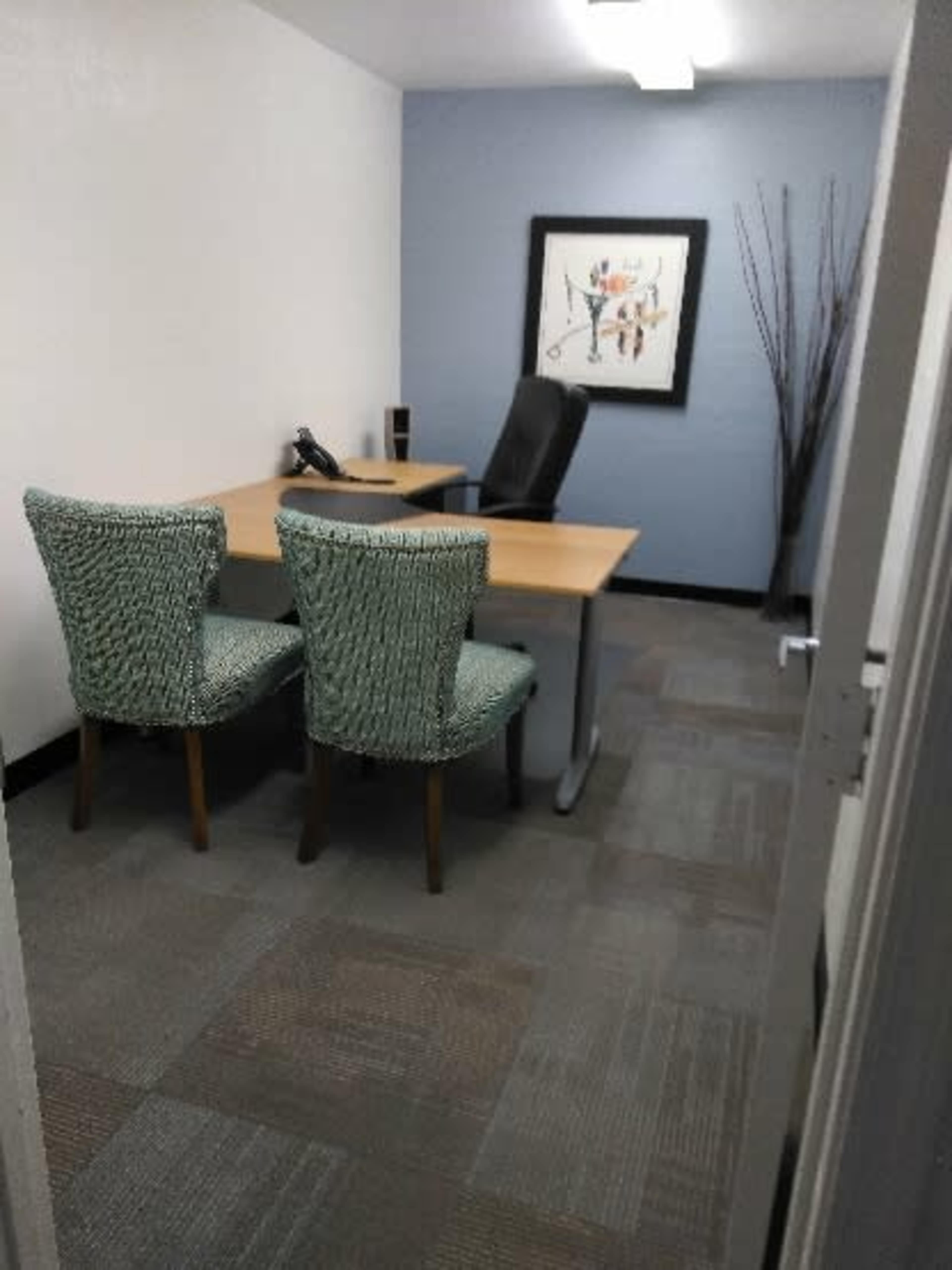 The image shows a small office space featuring a desk with a phone, a leather chair, and two green textured chairs positioned at the desk.