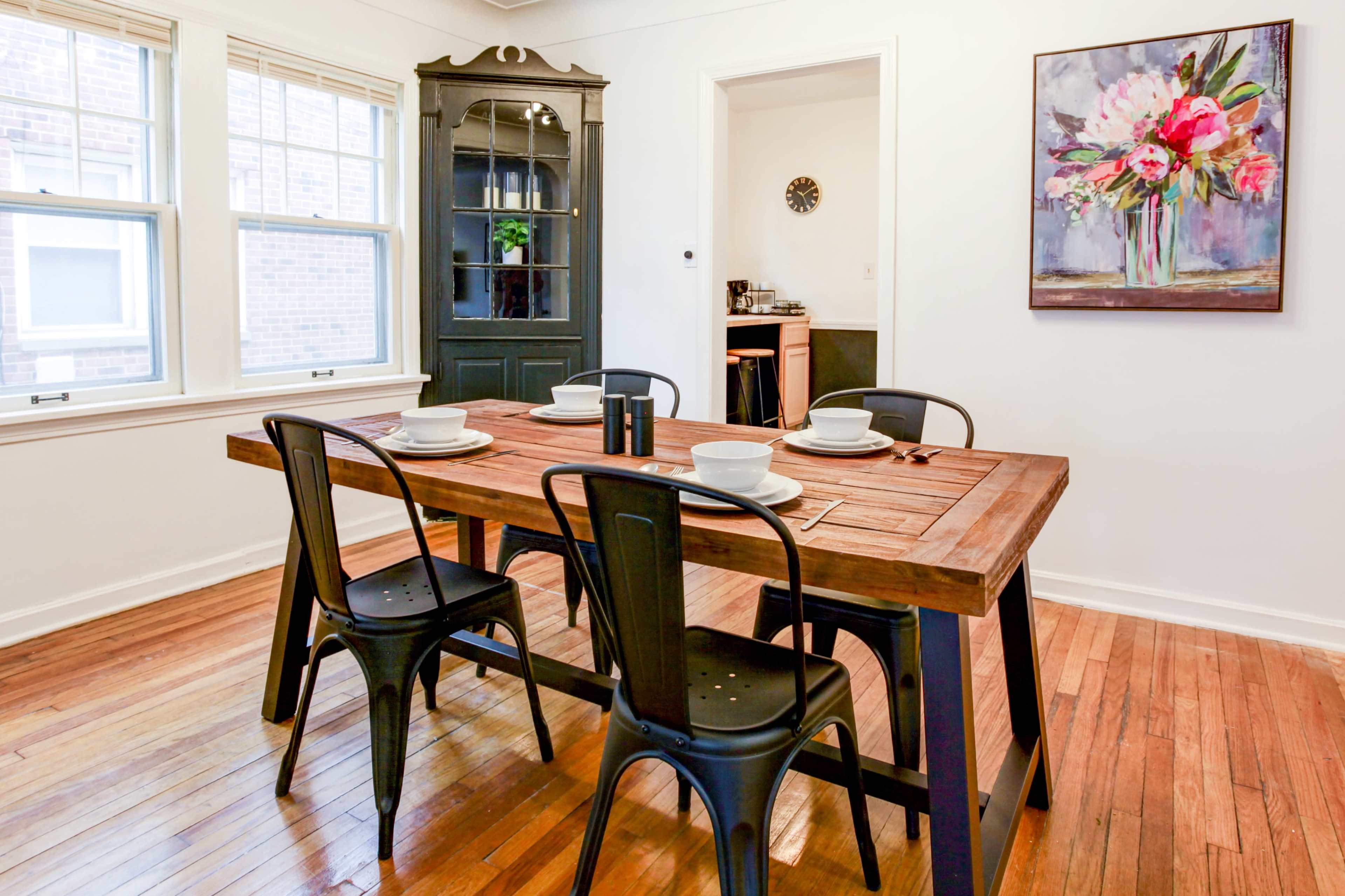 A wooden dining table with black chairs is set for four in a well-lit dining area featuring a black cabinet and a floral painting on the wall.