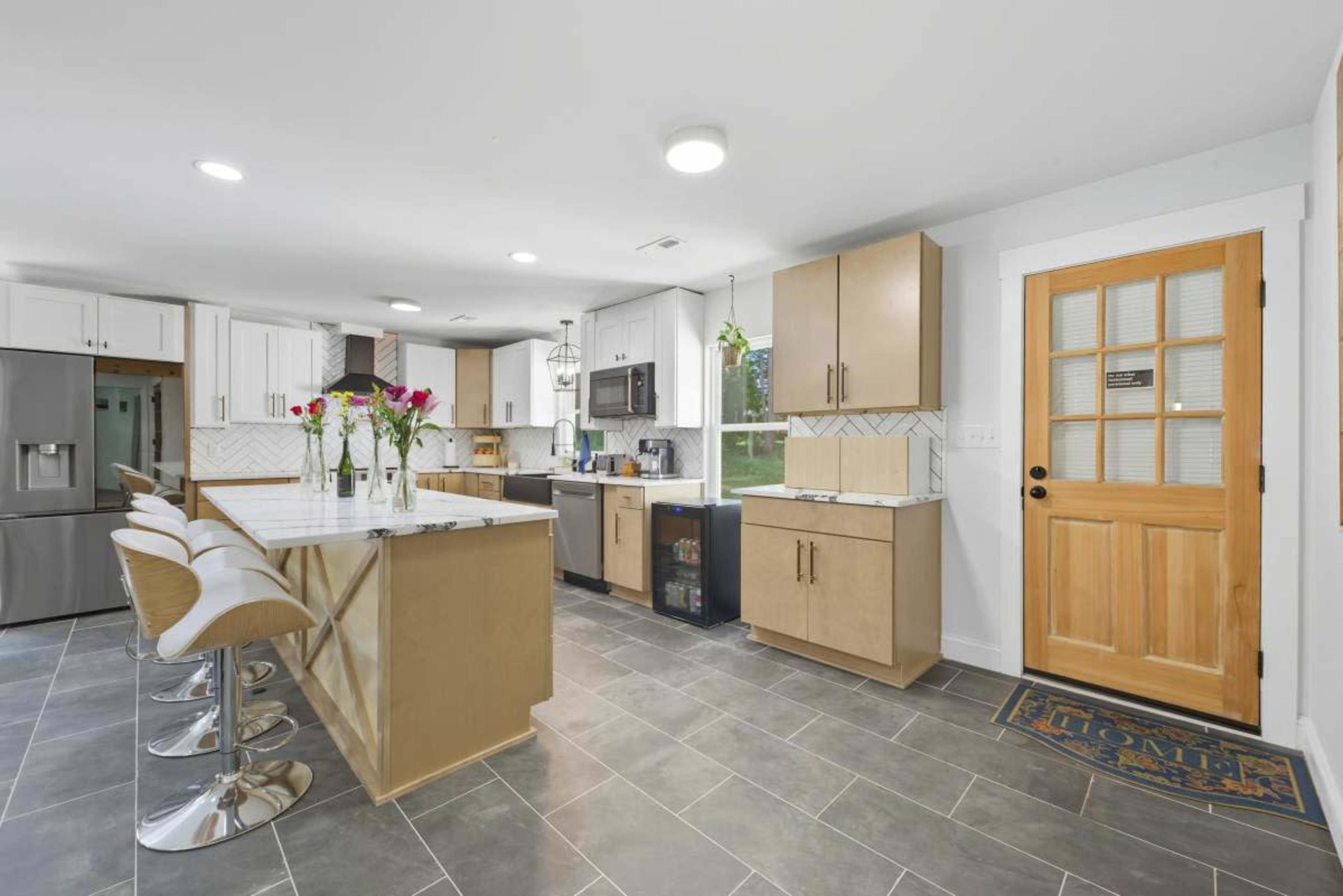 A modern kitchen features light wooden cabinets, a large island with seating, and sleek appliances, set against gray tile flooring.