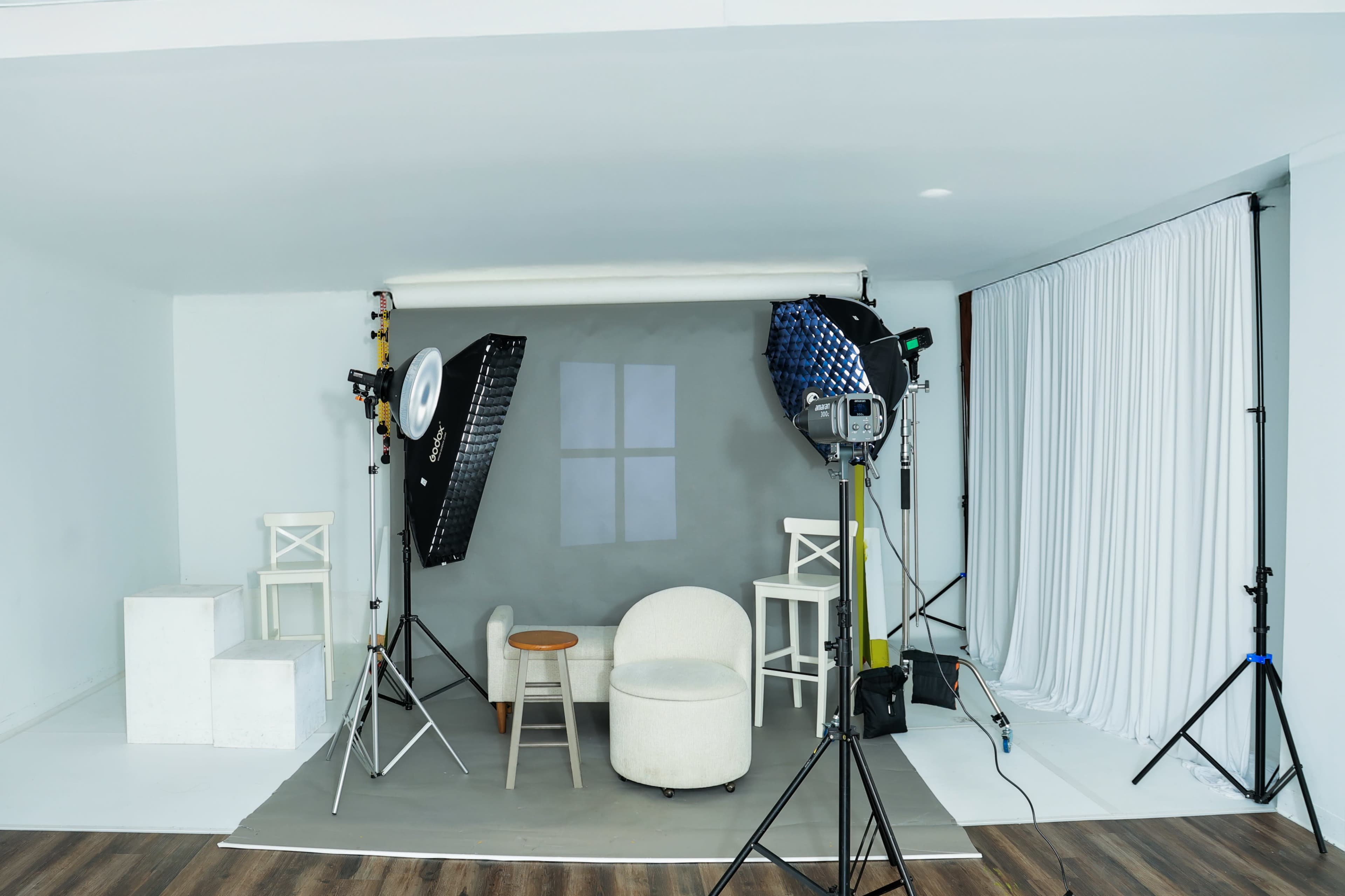 The image shows a photography studio setup featuring a gray backdrop, a chair, a stool, lighting equipment, and draped white fabric.