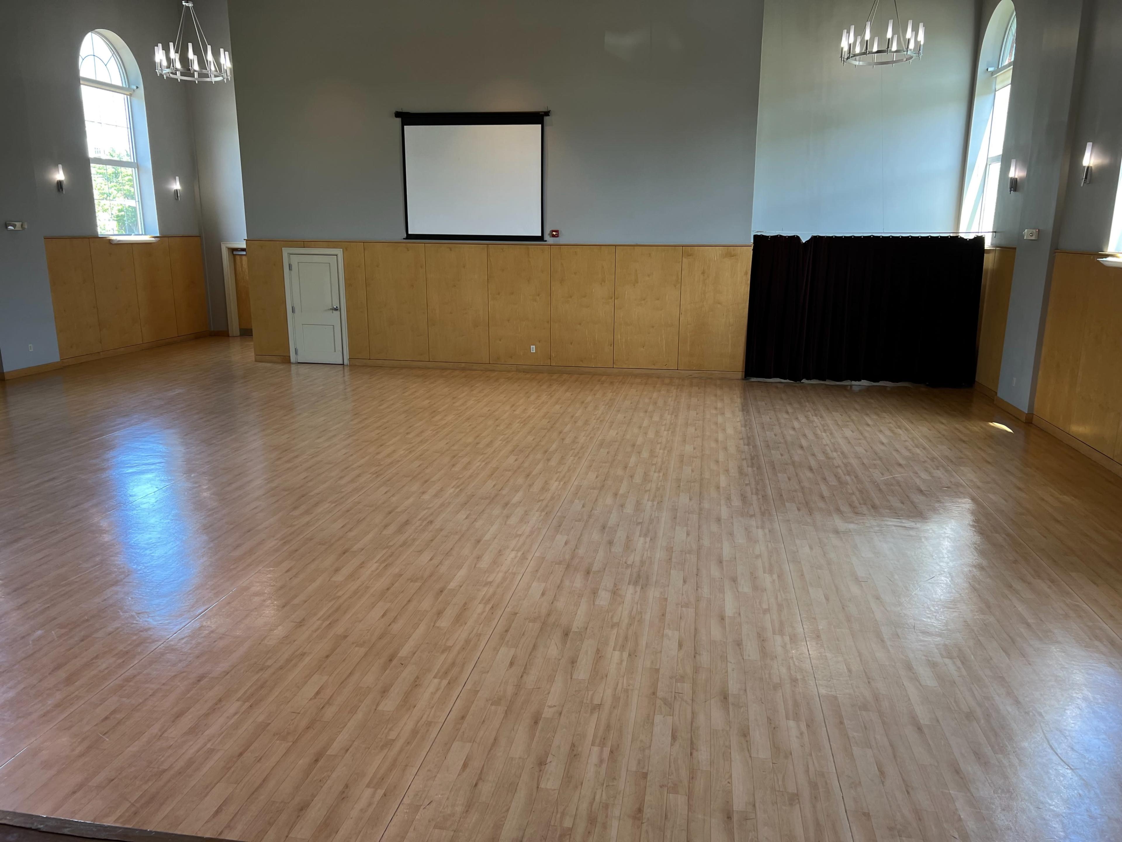 A spacious, empty hall with wooden paneling, a projection screen on the wall, and large windows allowing natural light.