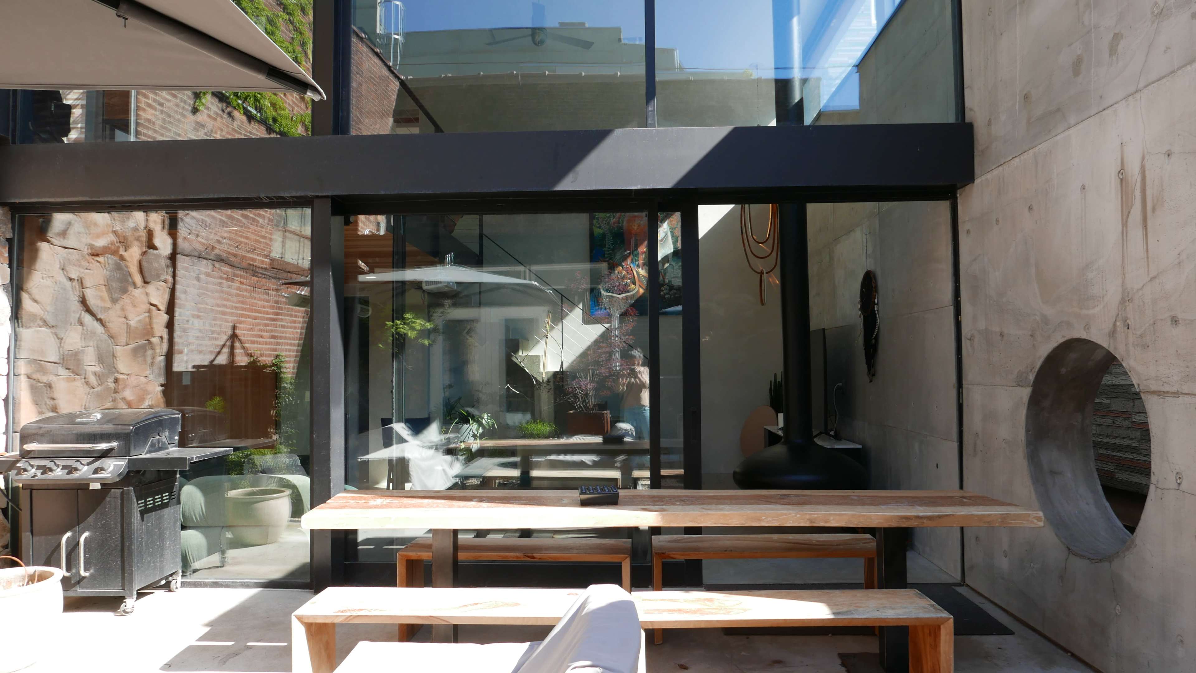 The image shows a modern outdoor patio with a wooden dining table and benches, framed by large glass walls that provide a view of the interior space.