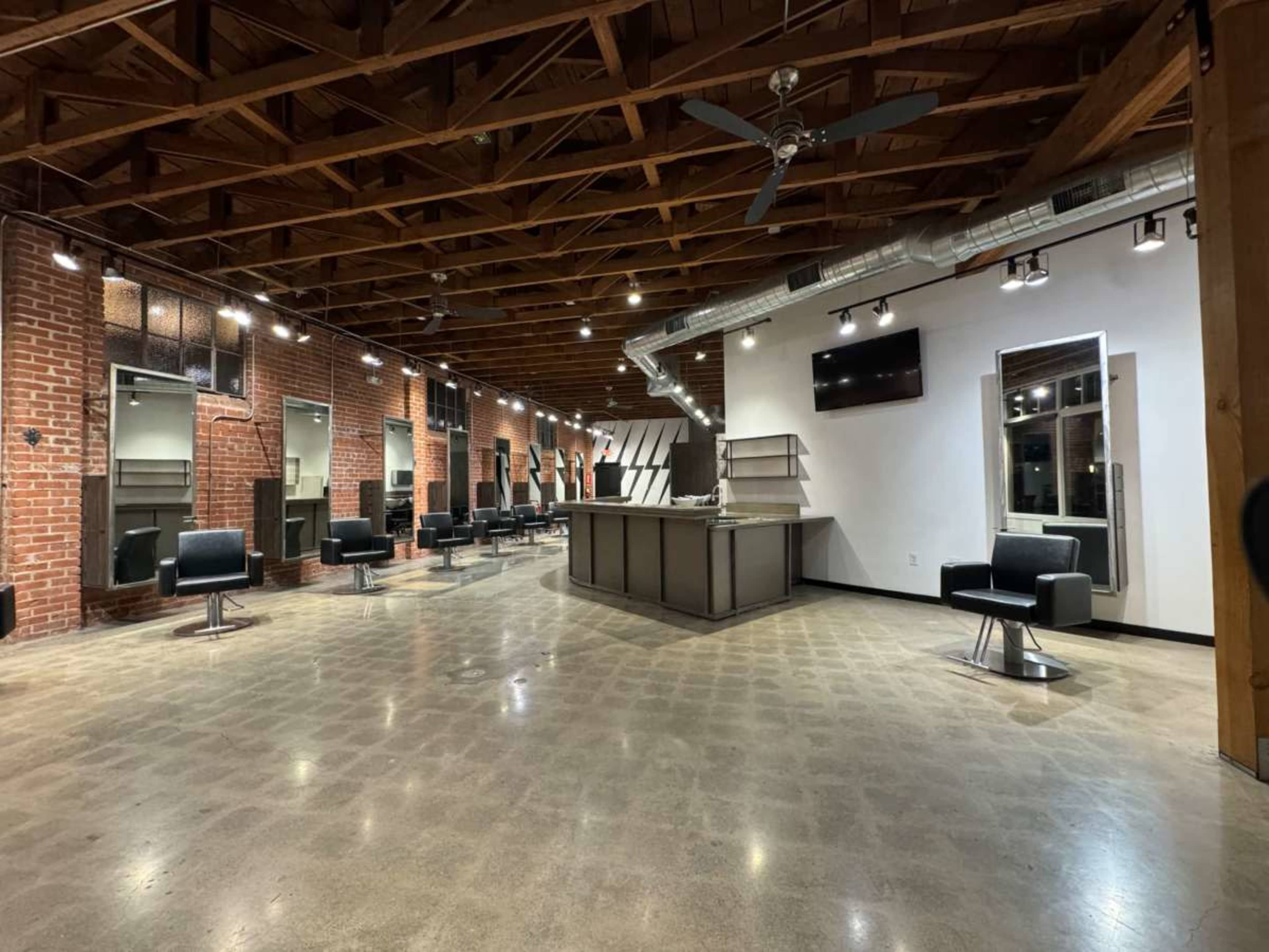 The image shows a spacious, modern salon with exposed brick walls, a central reception area, and several styling stations equipped with black chairs.
