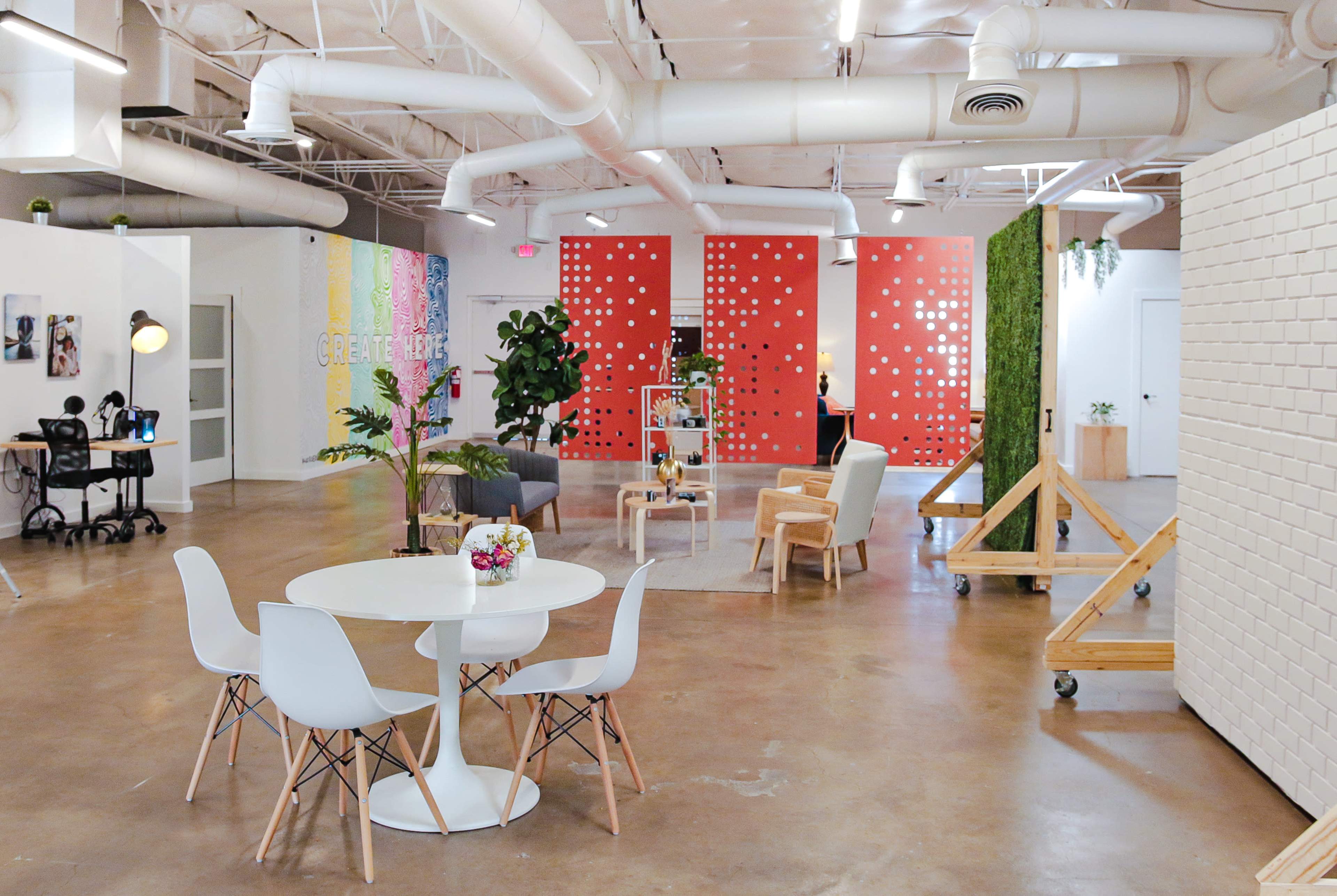 A modern workspace featuring a round table with white chairs, colorful accent walls, plants, and various seating areas.