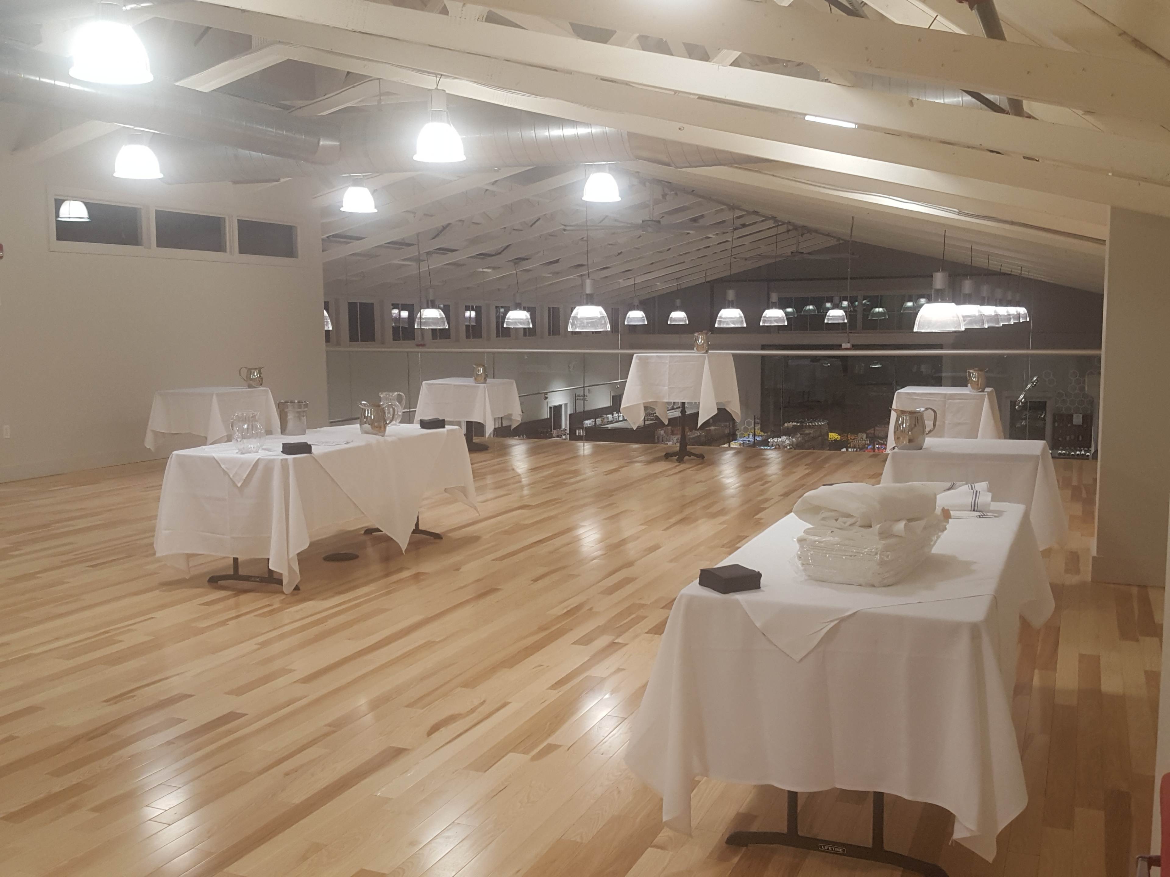 A spacious event venue with polished wooden floors and neatly arranged tables covered with white tablecloths under bright ceiling lights.