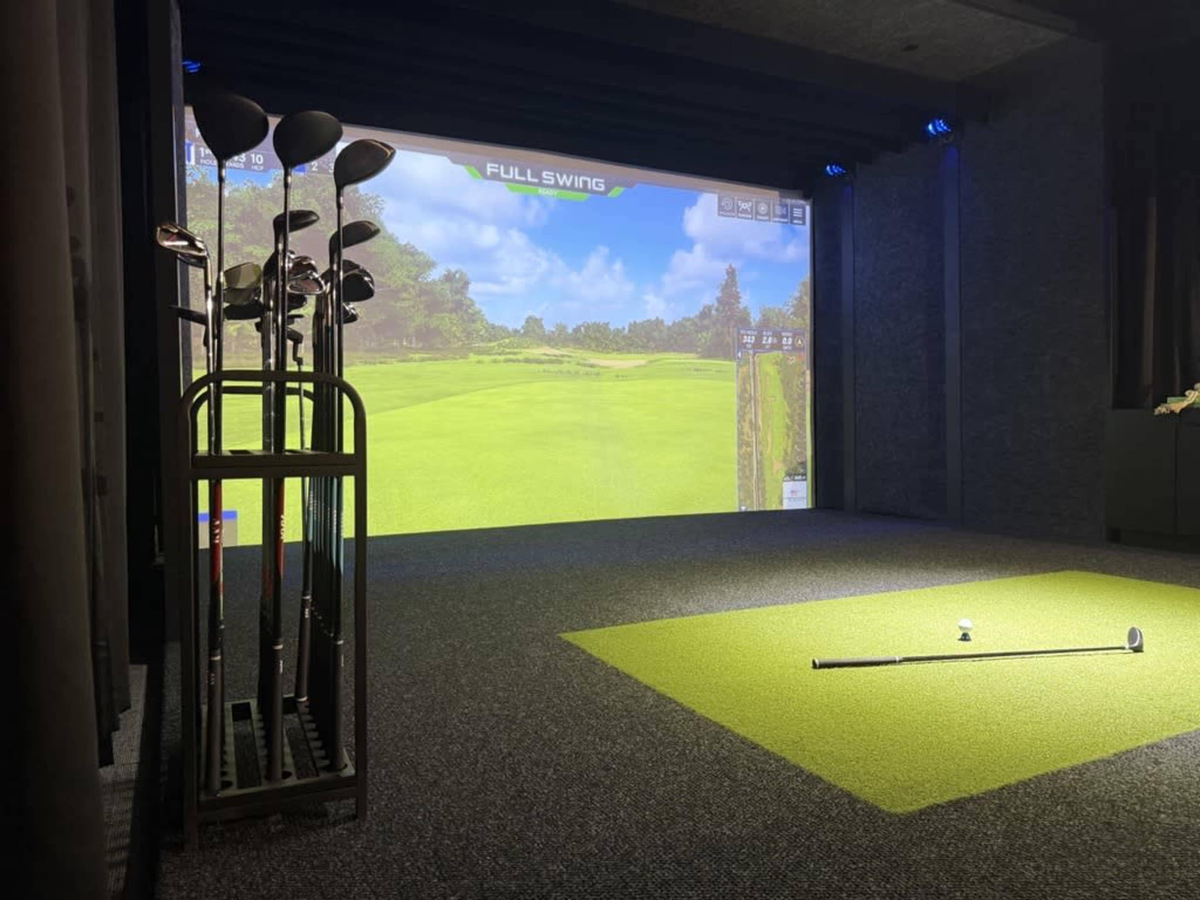 The image shows an indoor golf simulator setup with a screen displaying a green landscape and a golf tee with a club next to it.
