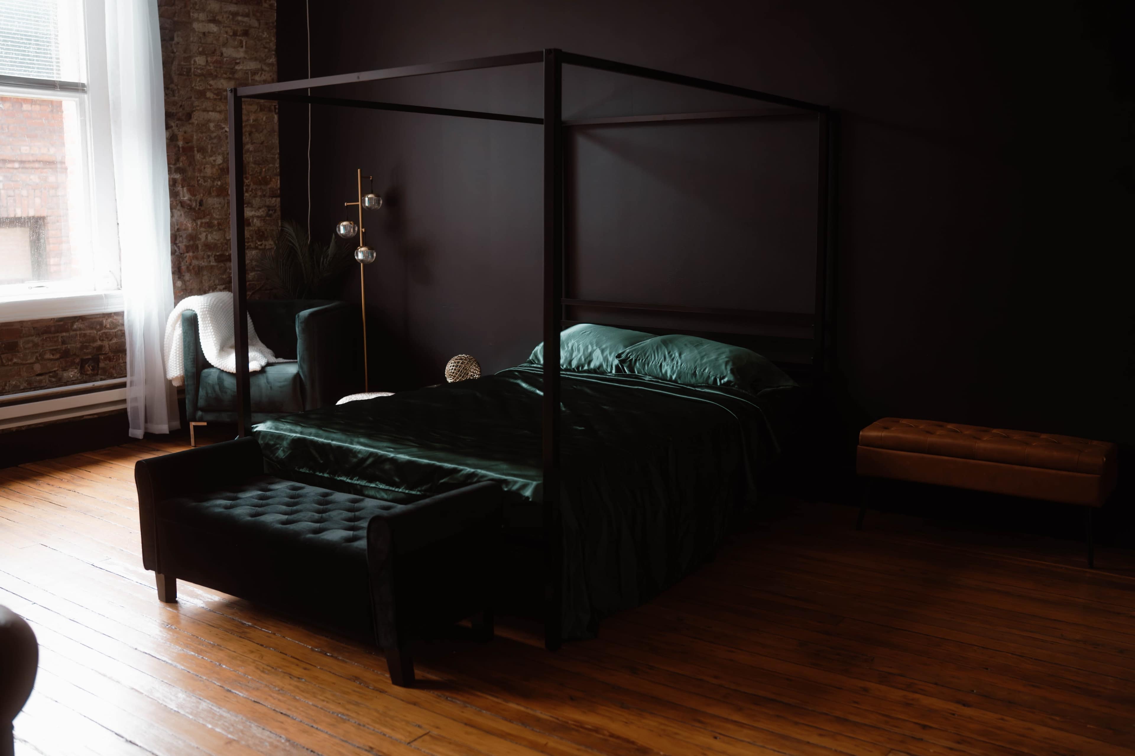 A four-poster bed with emerald green bedding is positioned in a dimly lit room featuring exposed brick walls and wooden flooring.