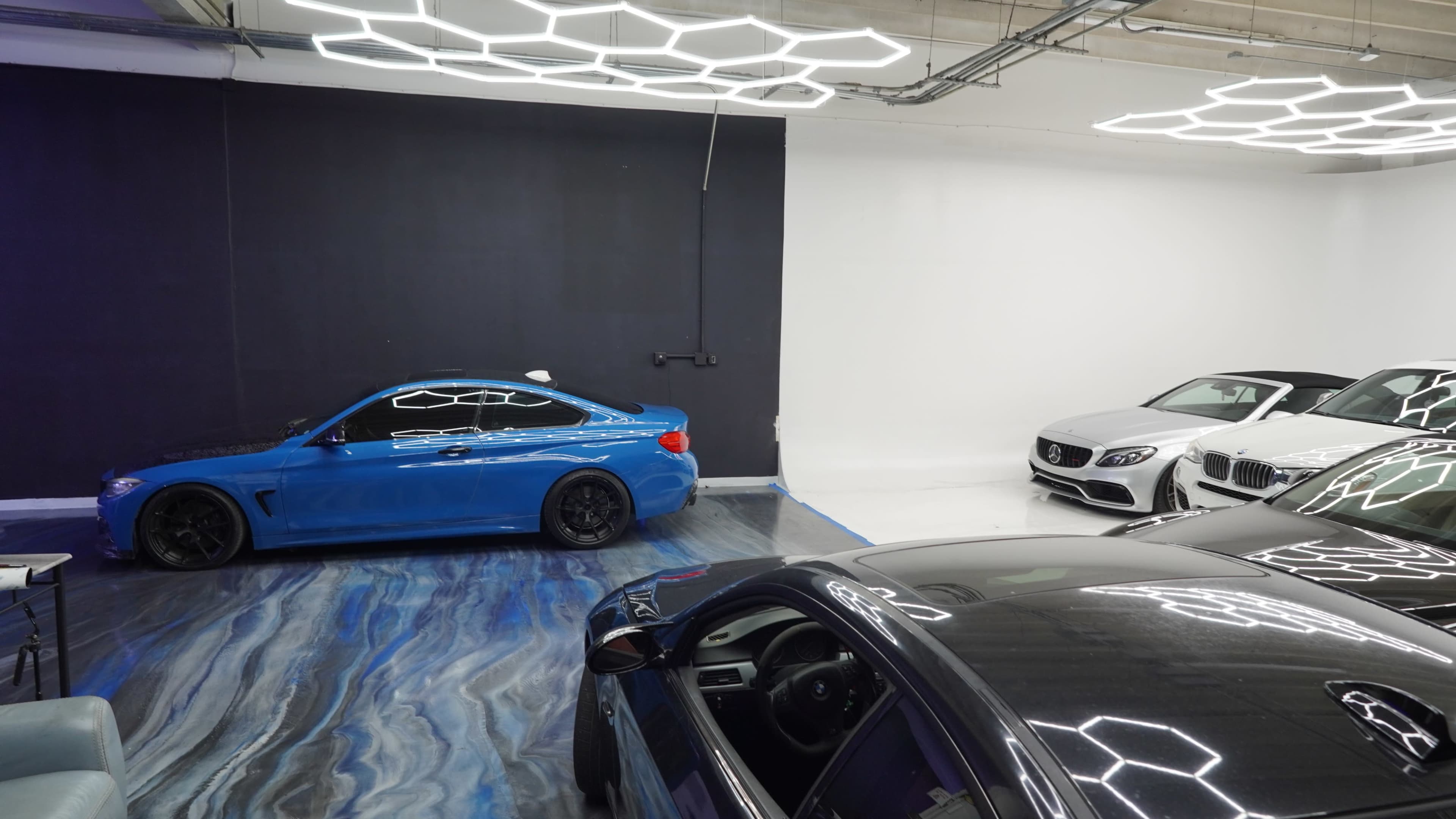 The image shows a modern car showroom featuring a blue sports car on the left and two luxury vehicles, a white sedan and a black car, on the right.