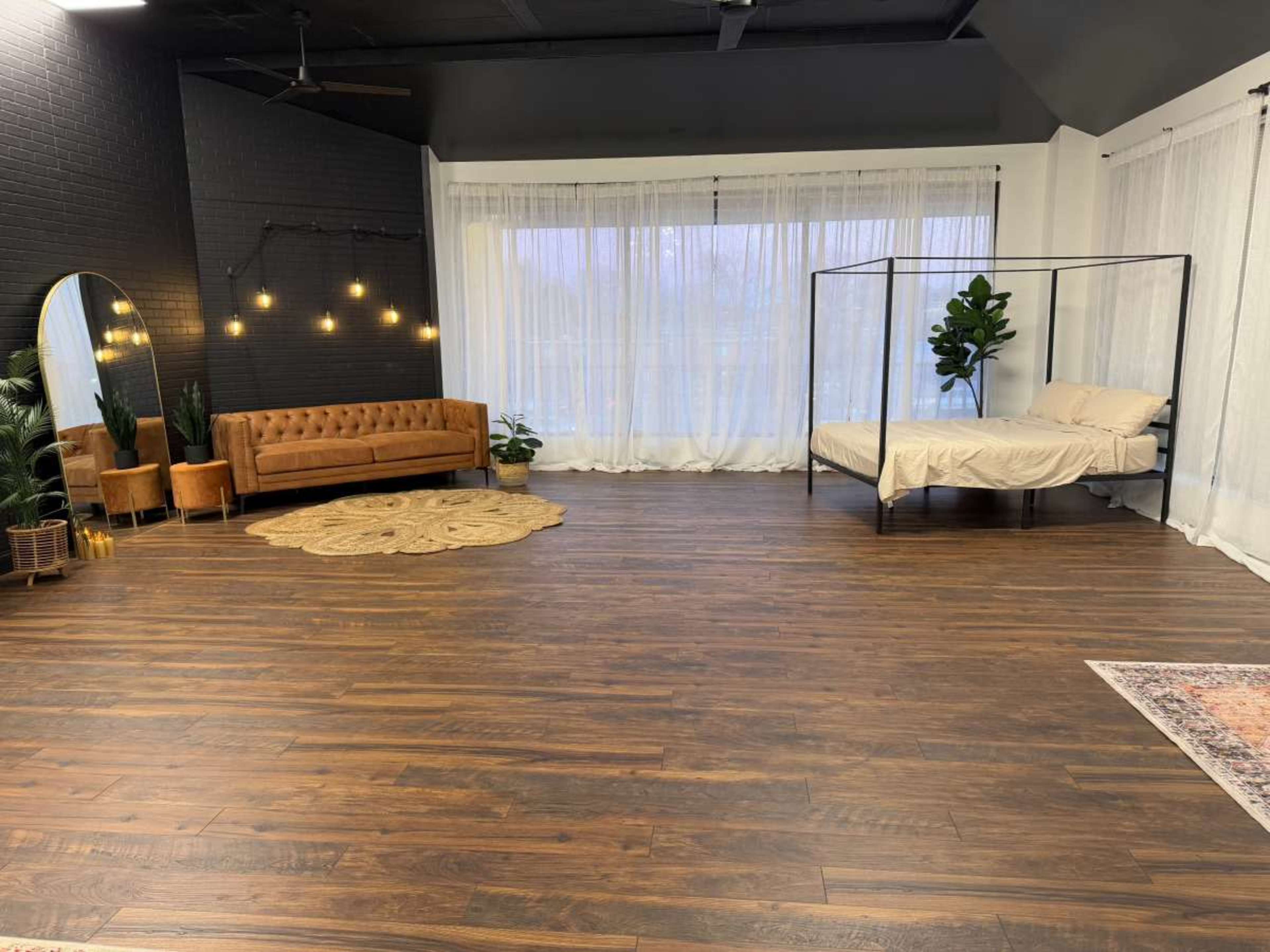 The image shows a spacious, modern indoor setting featuring a brown velvet couch, a bed with a canopy frame, large windows with sheer curtains, and decorative plants on a wooden floor.