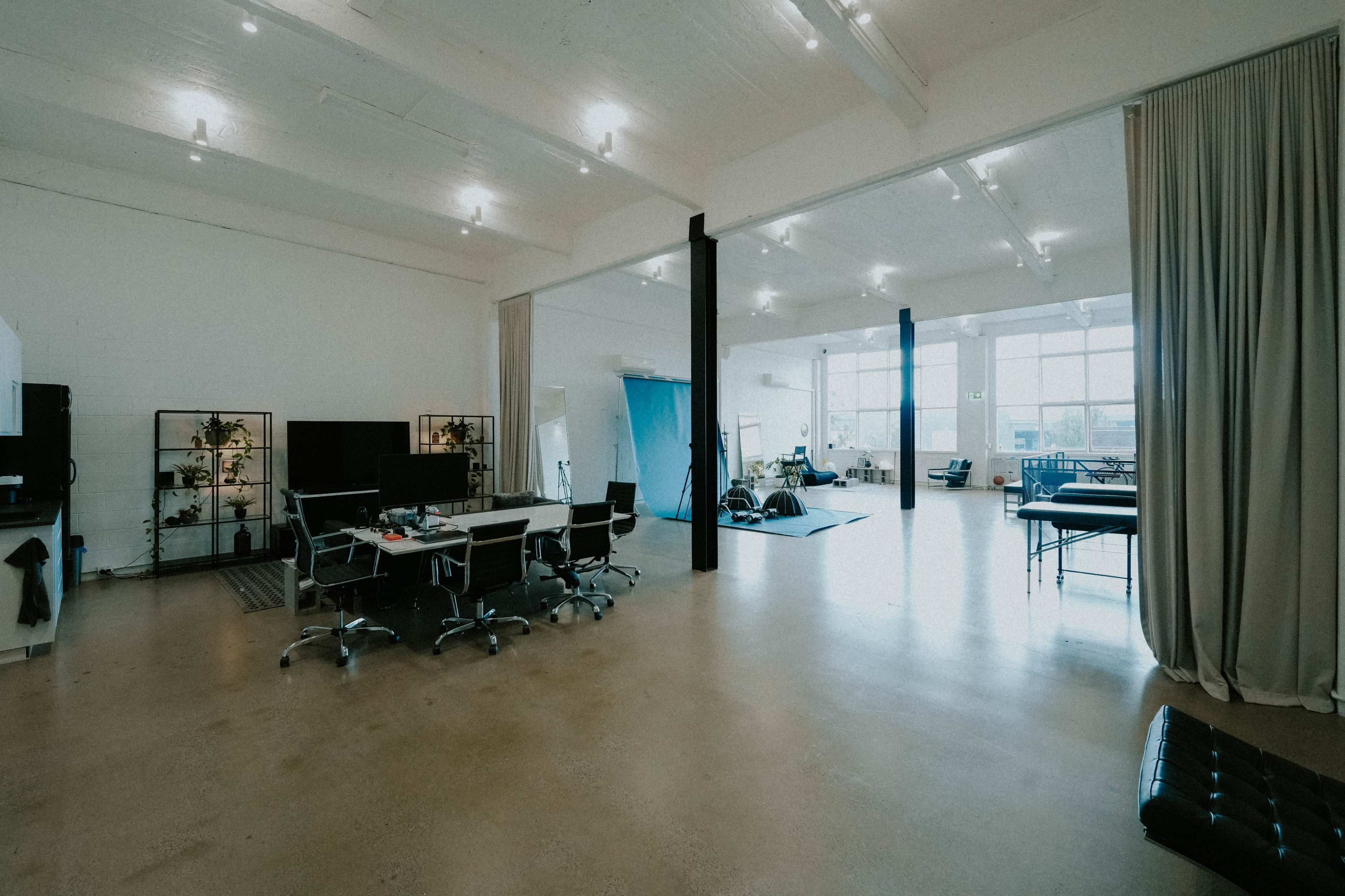 The image shows a spacious, modern office interior featuring a row of desks with chairs, large windows allowing natural light, and various seating areas in the background.