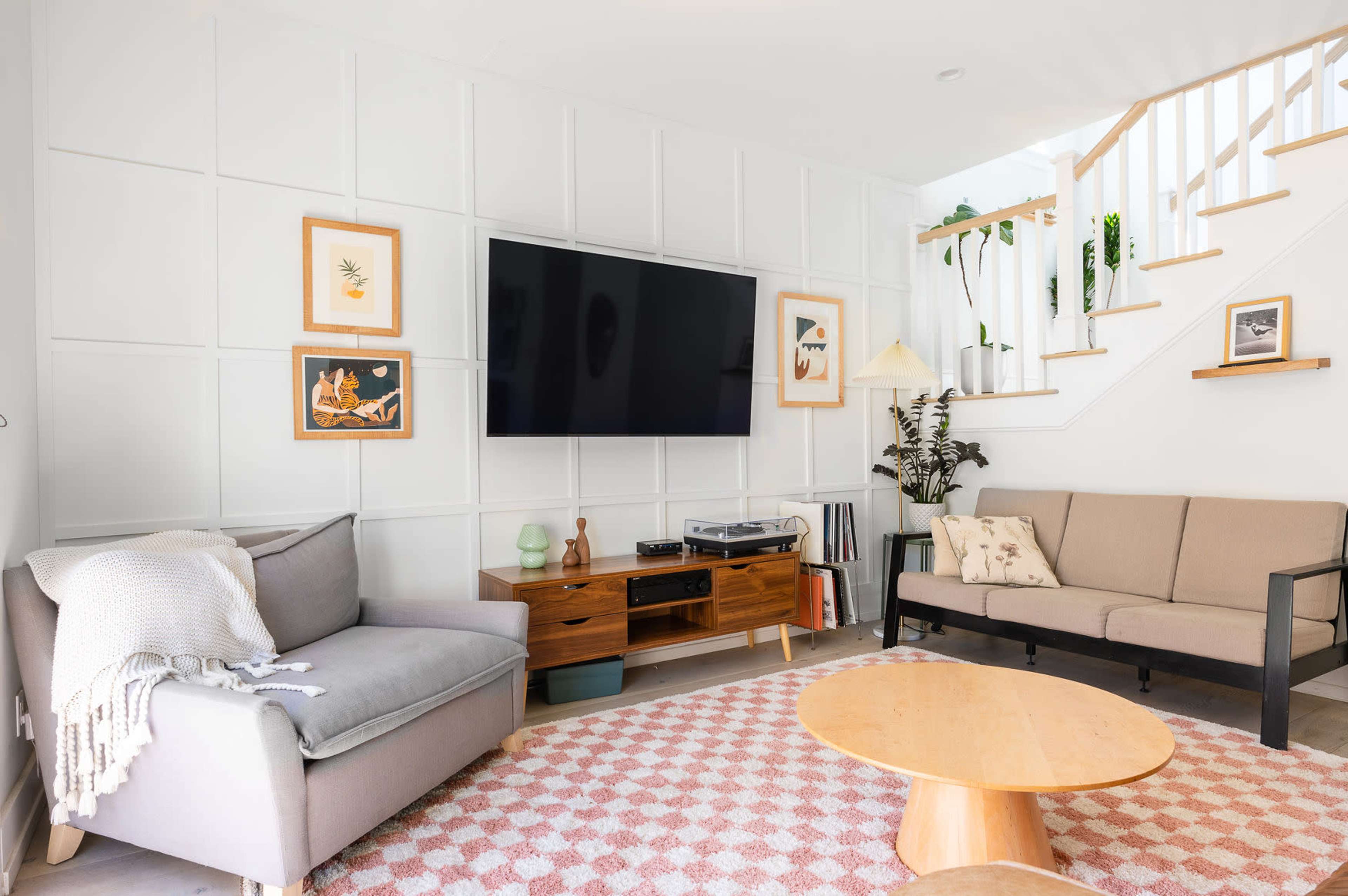 A living room with a television mounted on a textured wall, two sofas, a round coffee table, and stairs leading to an upper level.