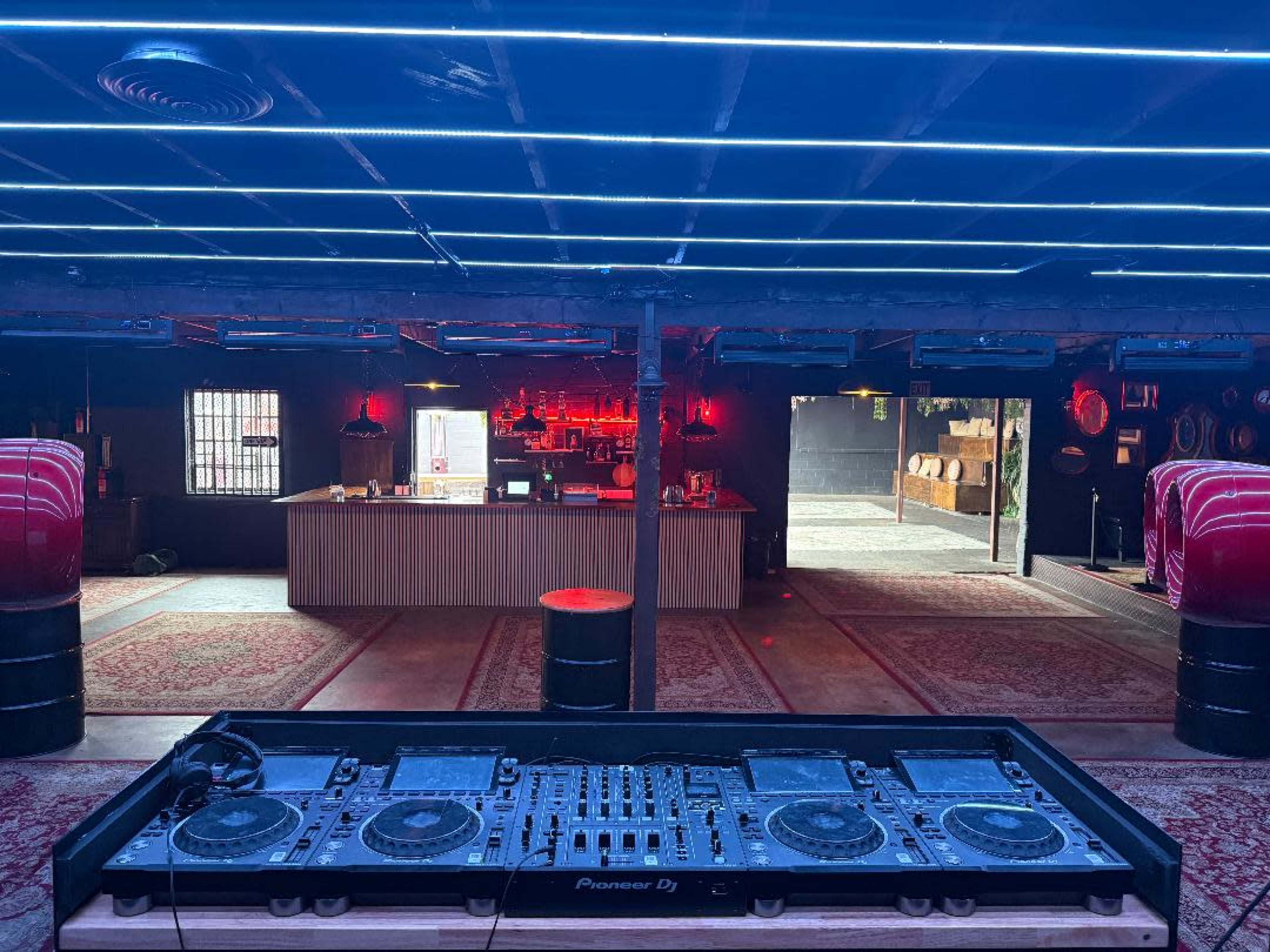 The image shows a DJ setup in a dimly lit venue with a bar in the background and a decorated floor.