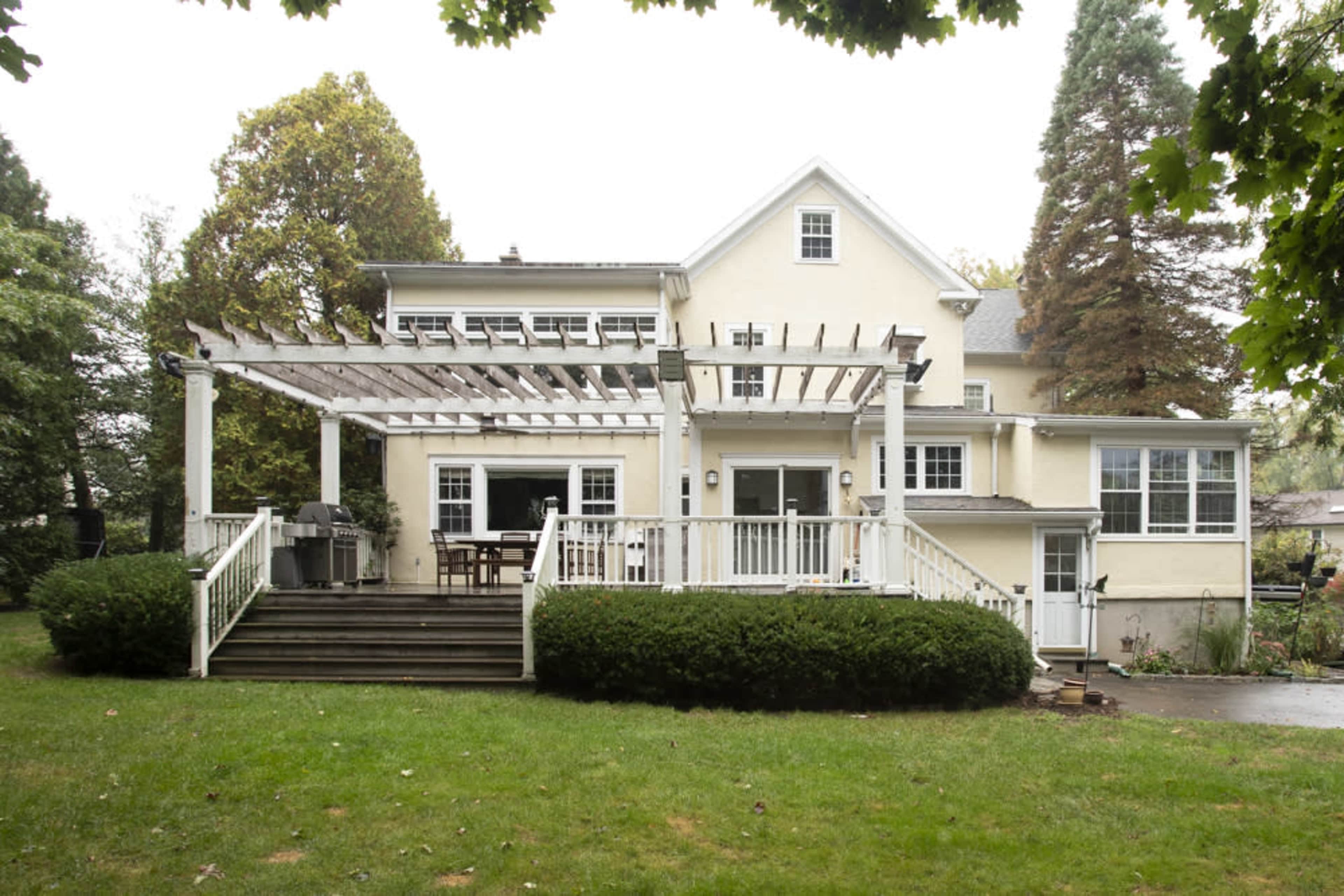 A two-story yellow house features a large wooden deck with a pergola, surrounded by green grass and shrubs.