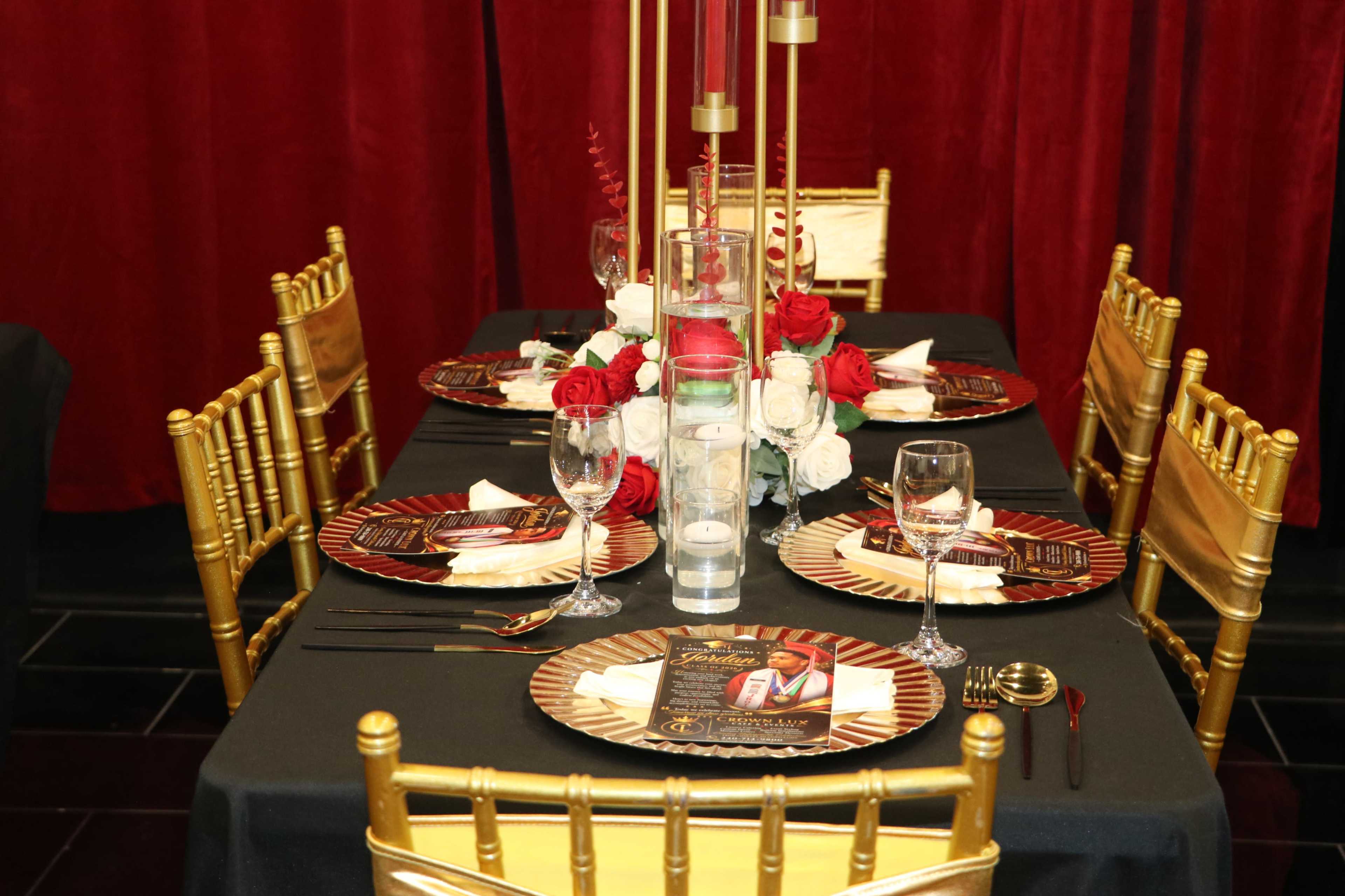 A banquet table is elegantly set with black tablecloths, gold chiavari chairs, decorative plates, and floral centerpieces featuring red and white roses.