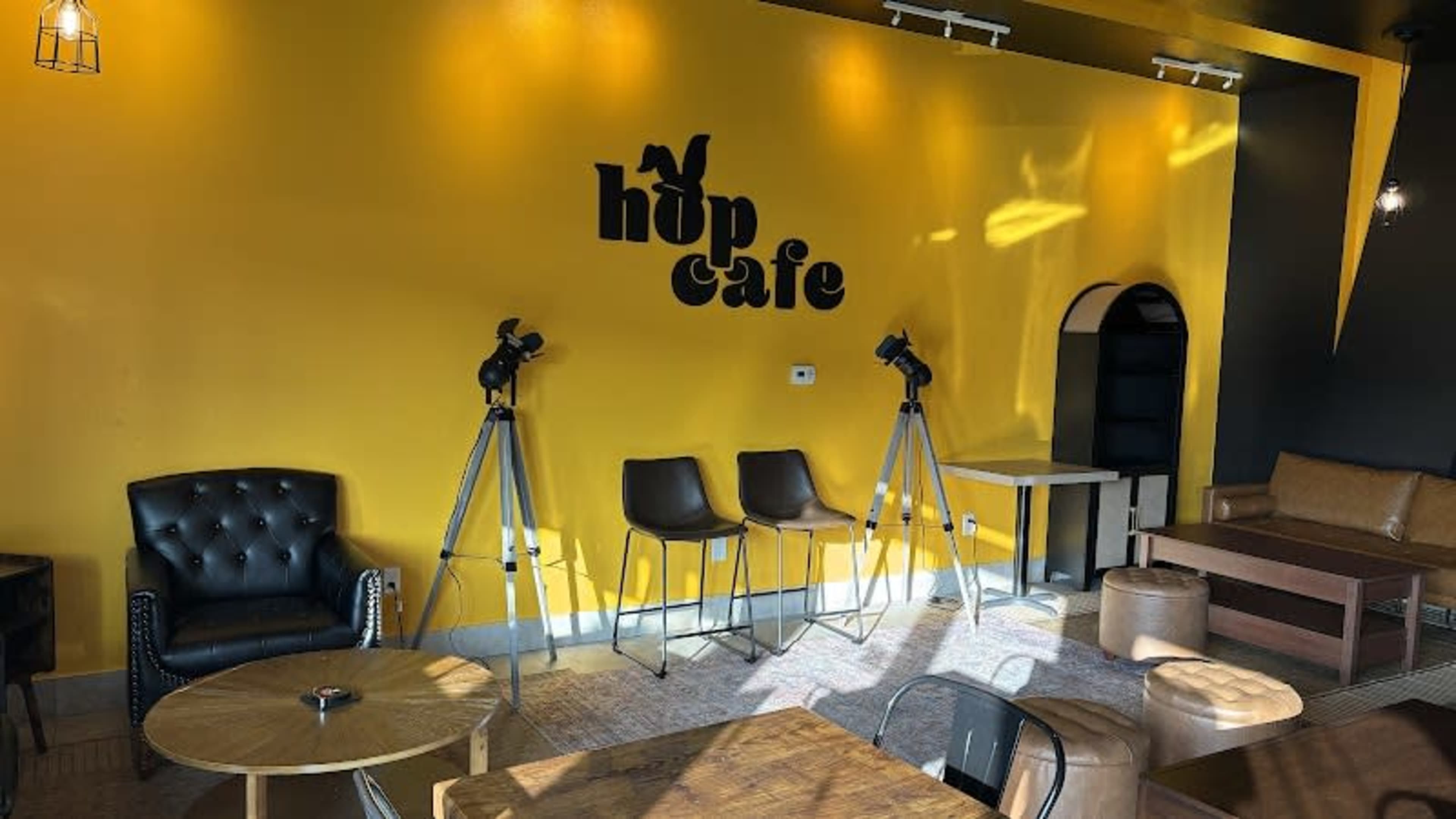 The image shows a cozy café interior with yellow walls, black furniture, and two cameras mounted on tripods.