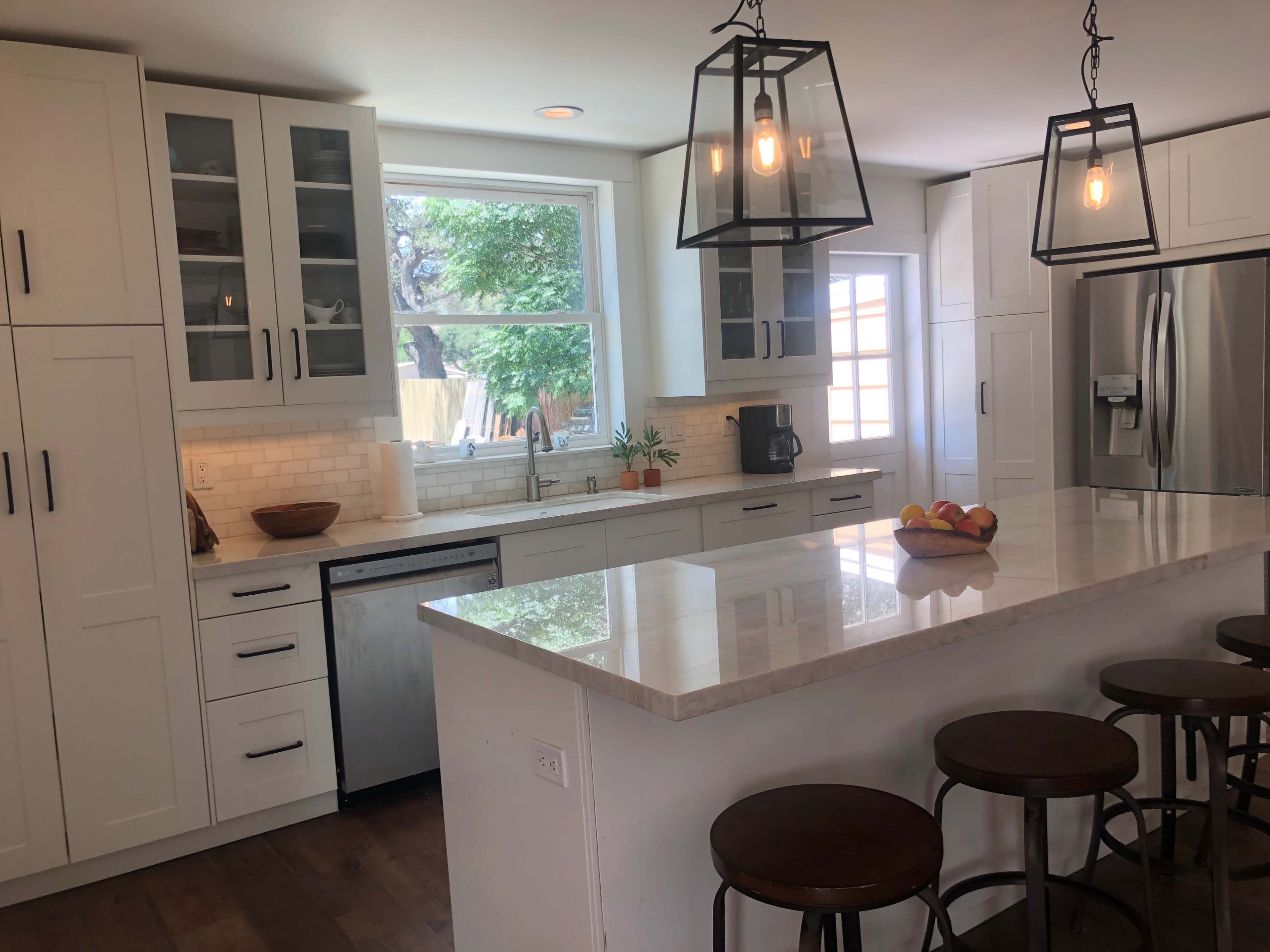 A modern kitchen features white cabinetry, a large island with bar stools, and large windows allowing natural light.