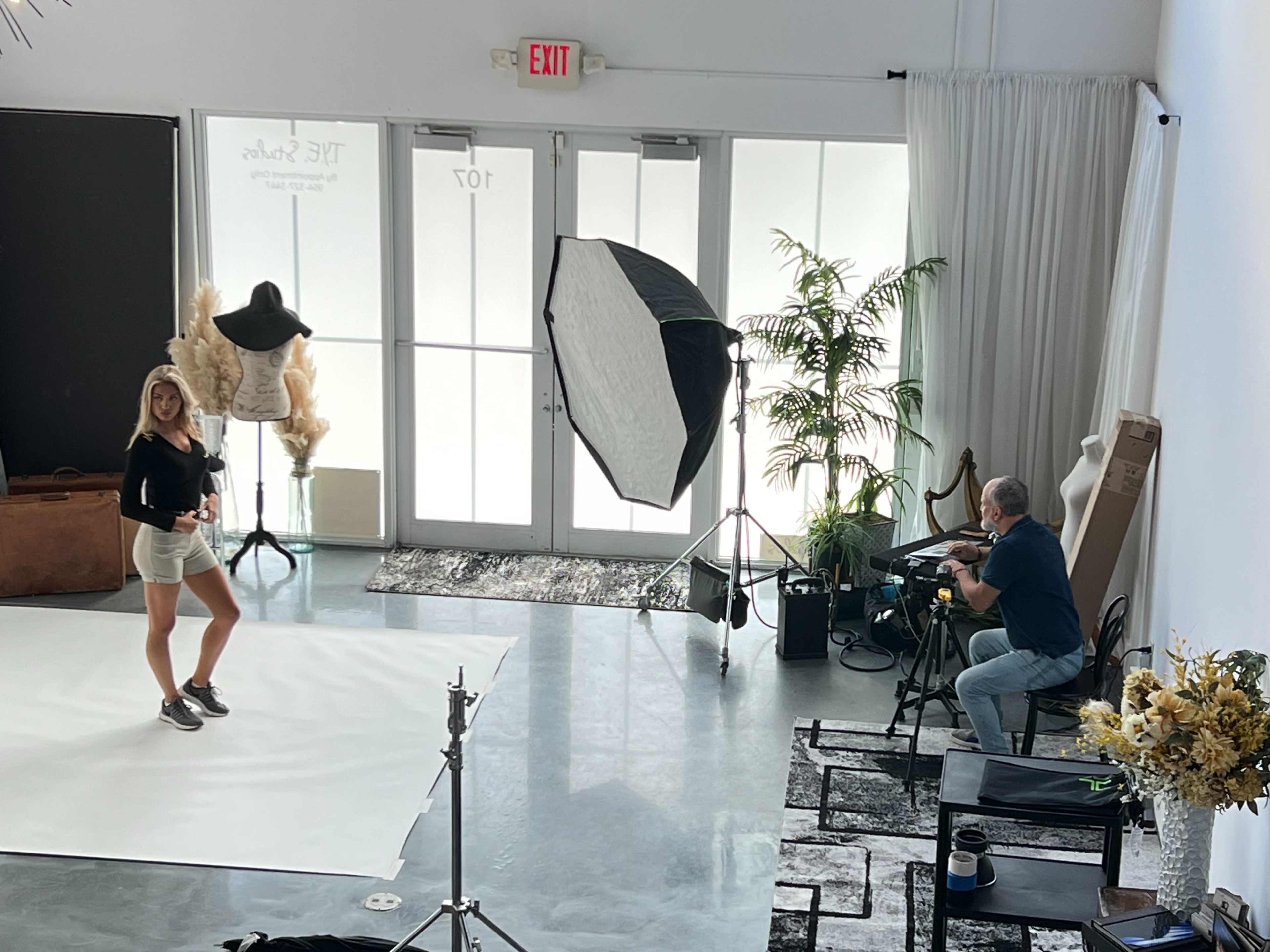 A model poses on a white backdrop while a photographer captures images from behind a camera in a studio setting.