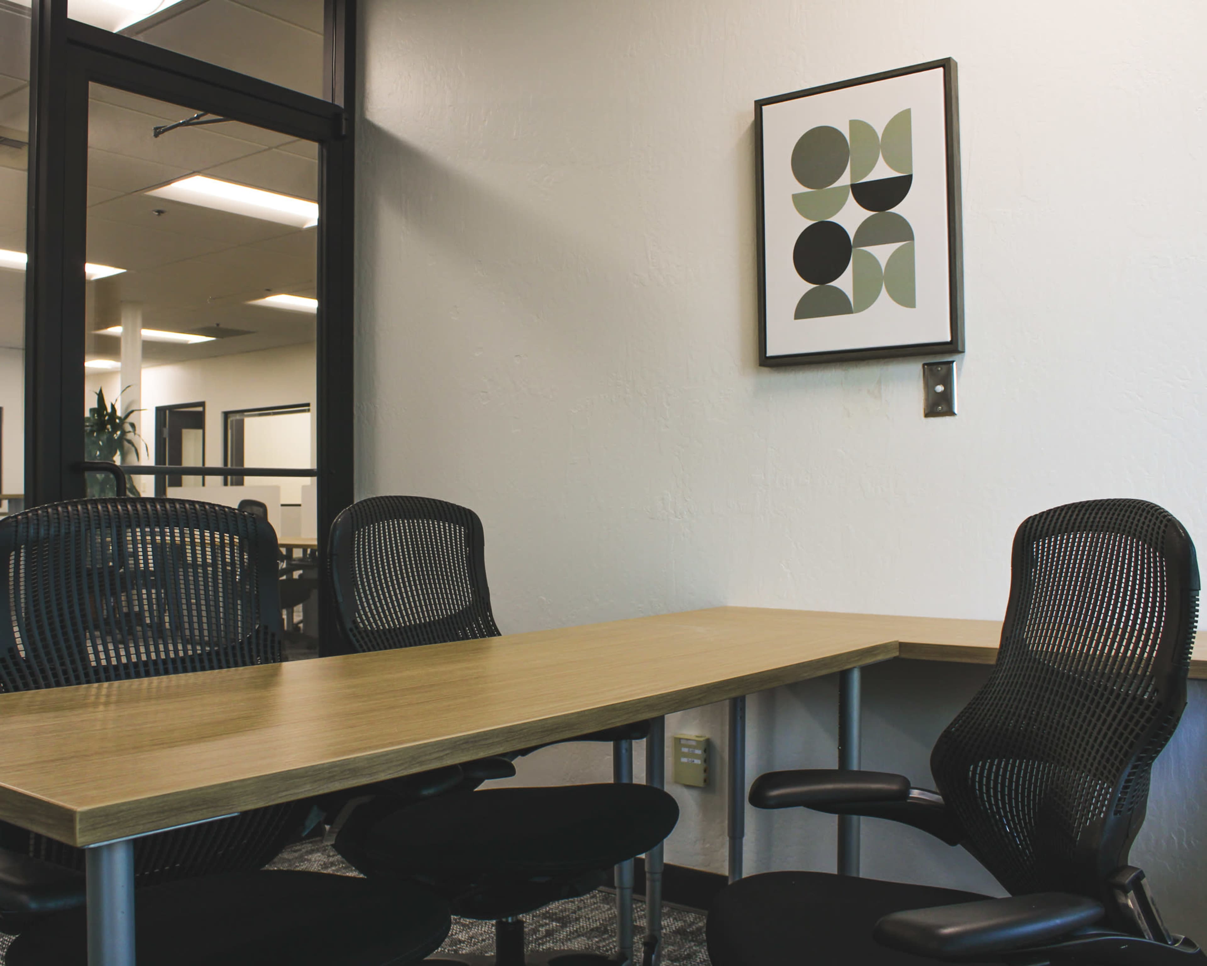 A simple office space features a wooden table and three ergonomic black chairs, with a framed abstract artwork on the wall.