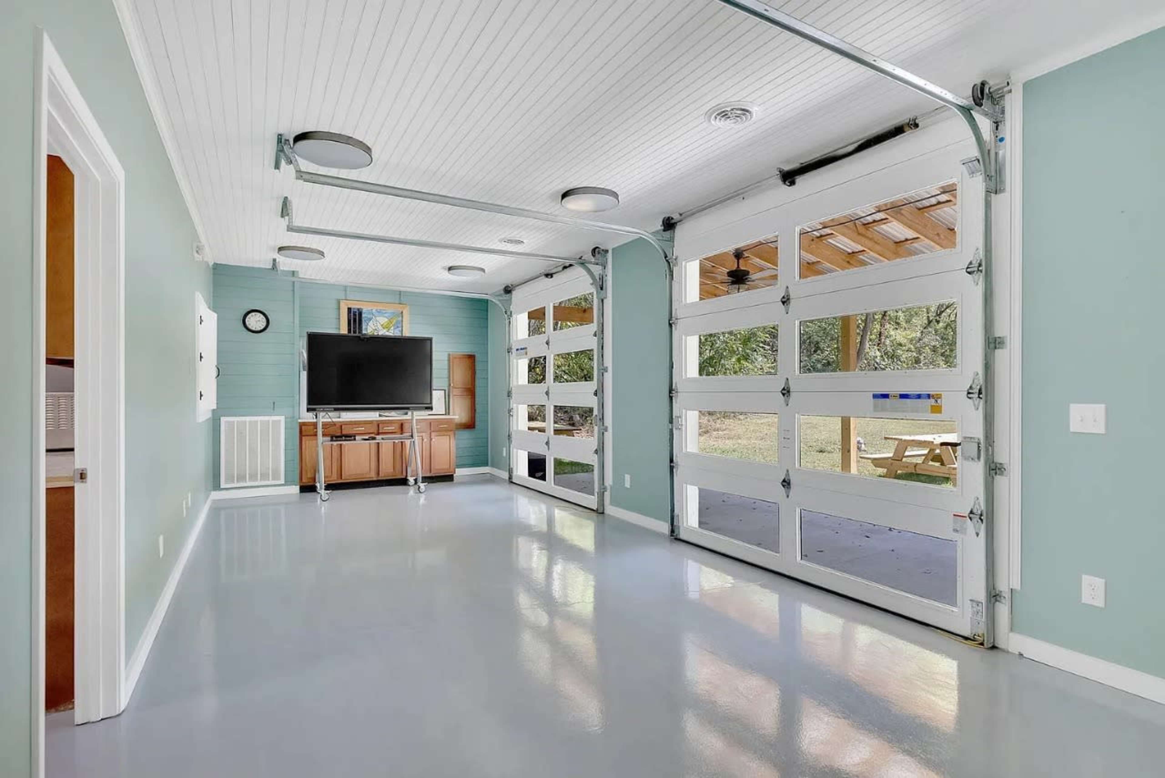 The image shows a clean garage space with large glass-paneled garage doors, a television mounted on a wall, and light blue walls adorned with minimal decor.