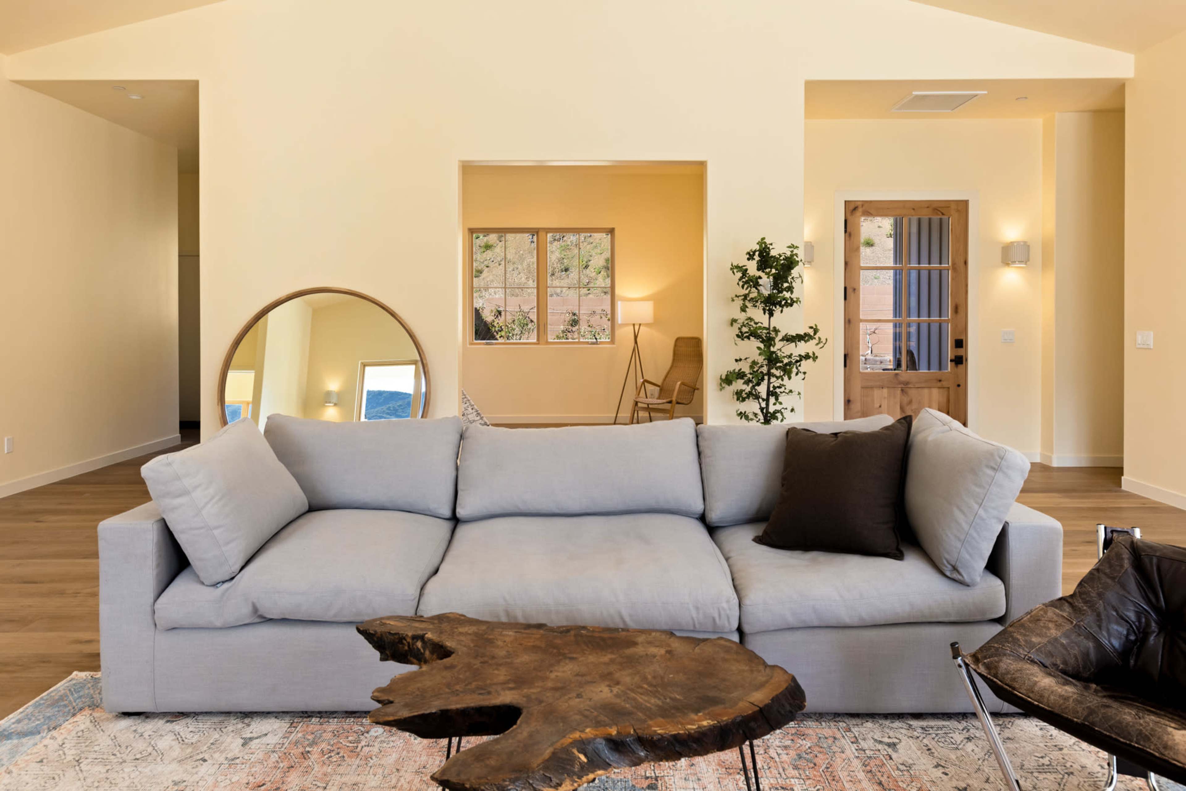 A modern living room with a light-colored sofa, a wooden coffee table, and a view of an adjoining room with a chair and window.