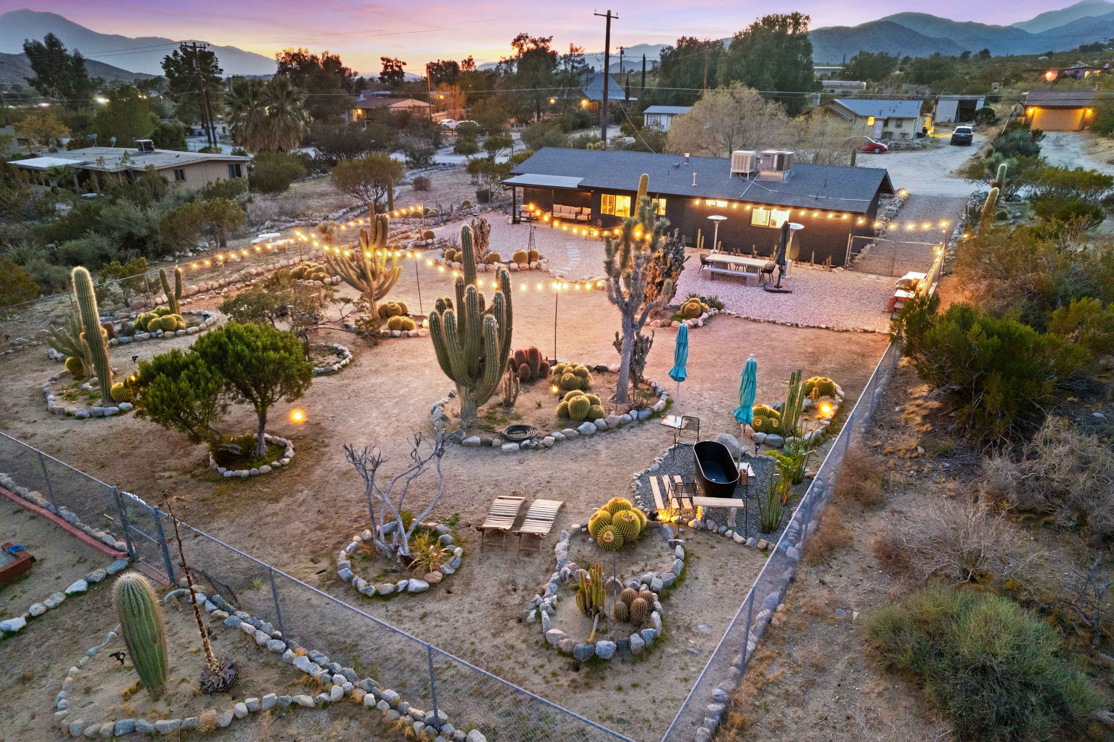 A desert property features a landscaped yard with cacti, seating areas, and strings of lights, surrounded by a fence and mountains in the background.