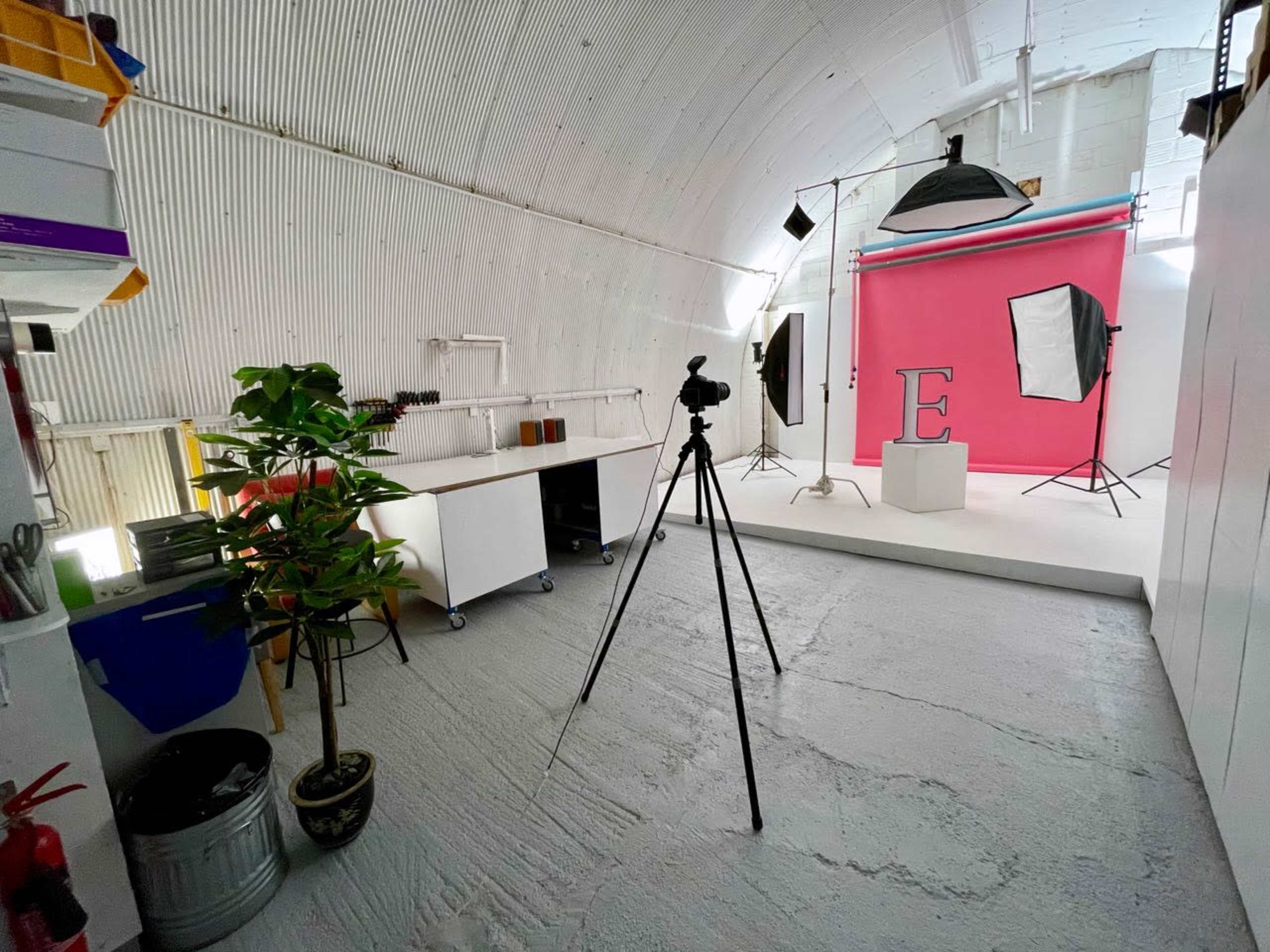 A photography studio featuring a bright pink backdrop, lighting equipment, and a tripod set up in front of a pedestal with the letter "E" displayed on it.