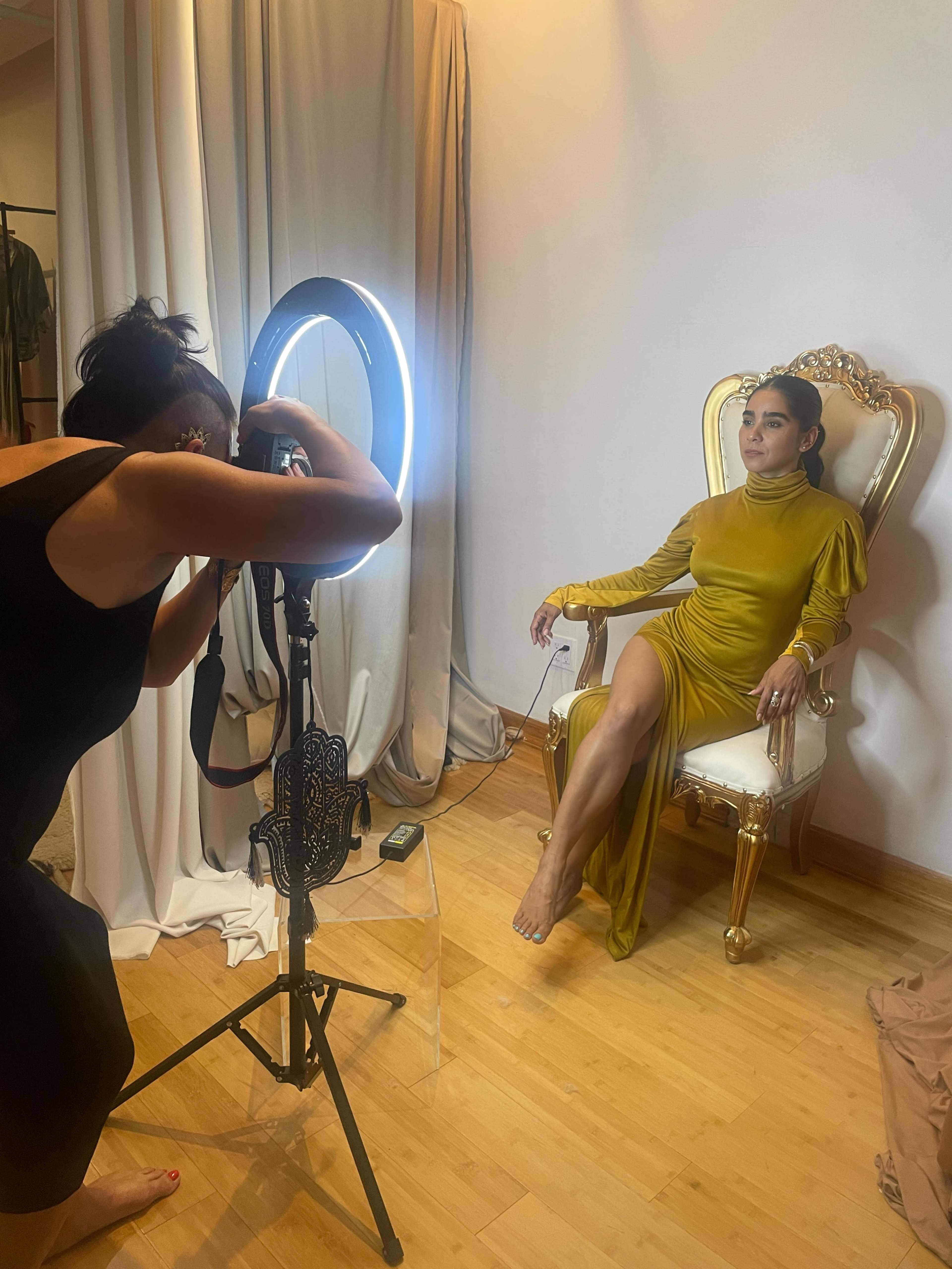 A photographer captures a model posing in a gold-accented chair while illuminated by a ring light.